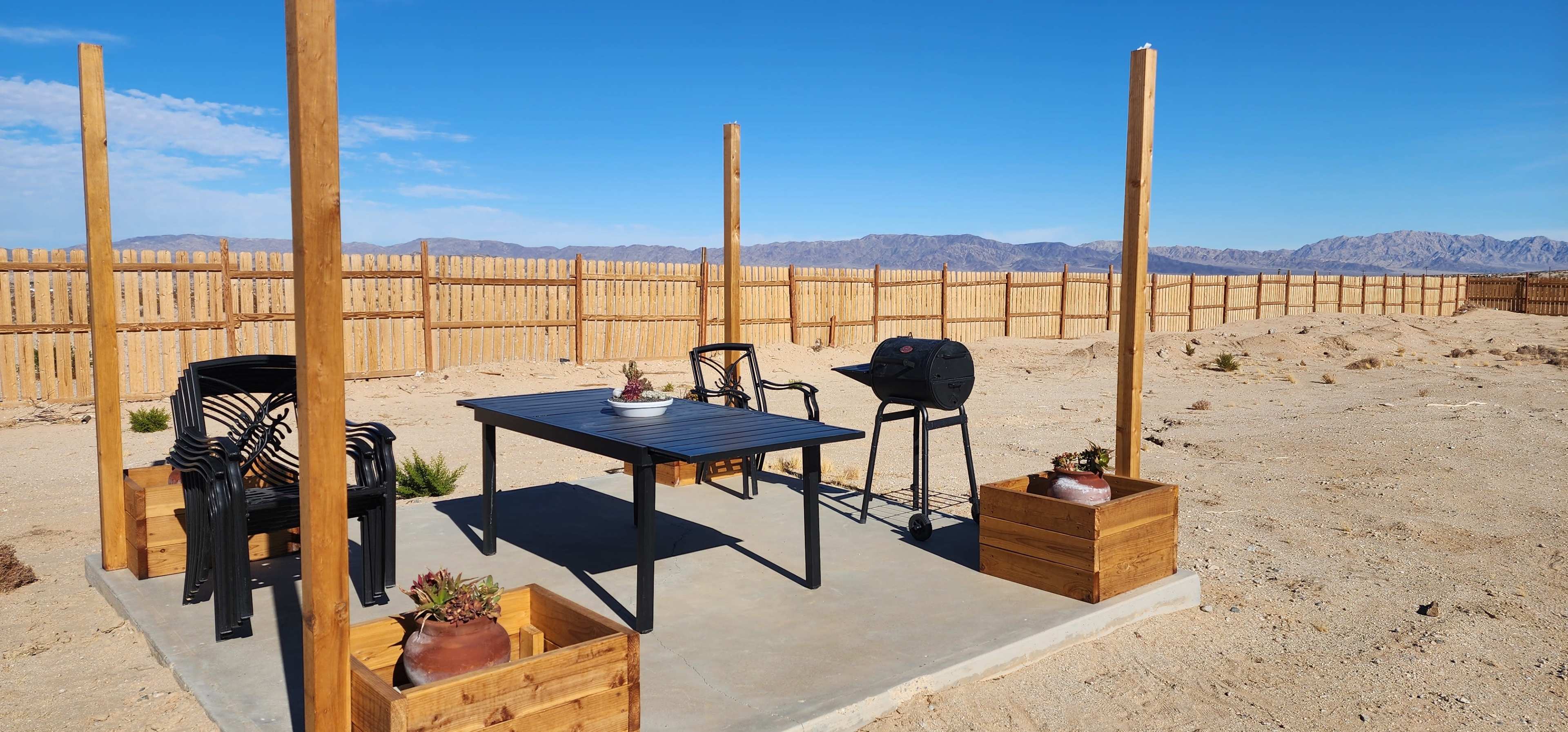 The image shows a simple outdoor seating area with a black table, two chairs, a grill, and potted plants, set in a sandy landscape with a wooden fence and mountains in the background.
