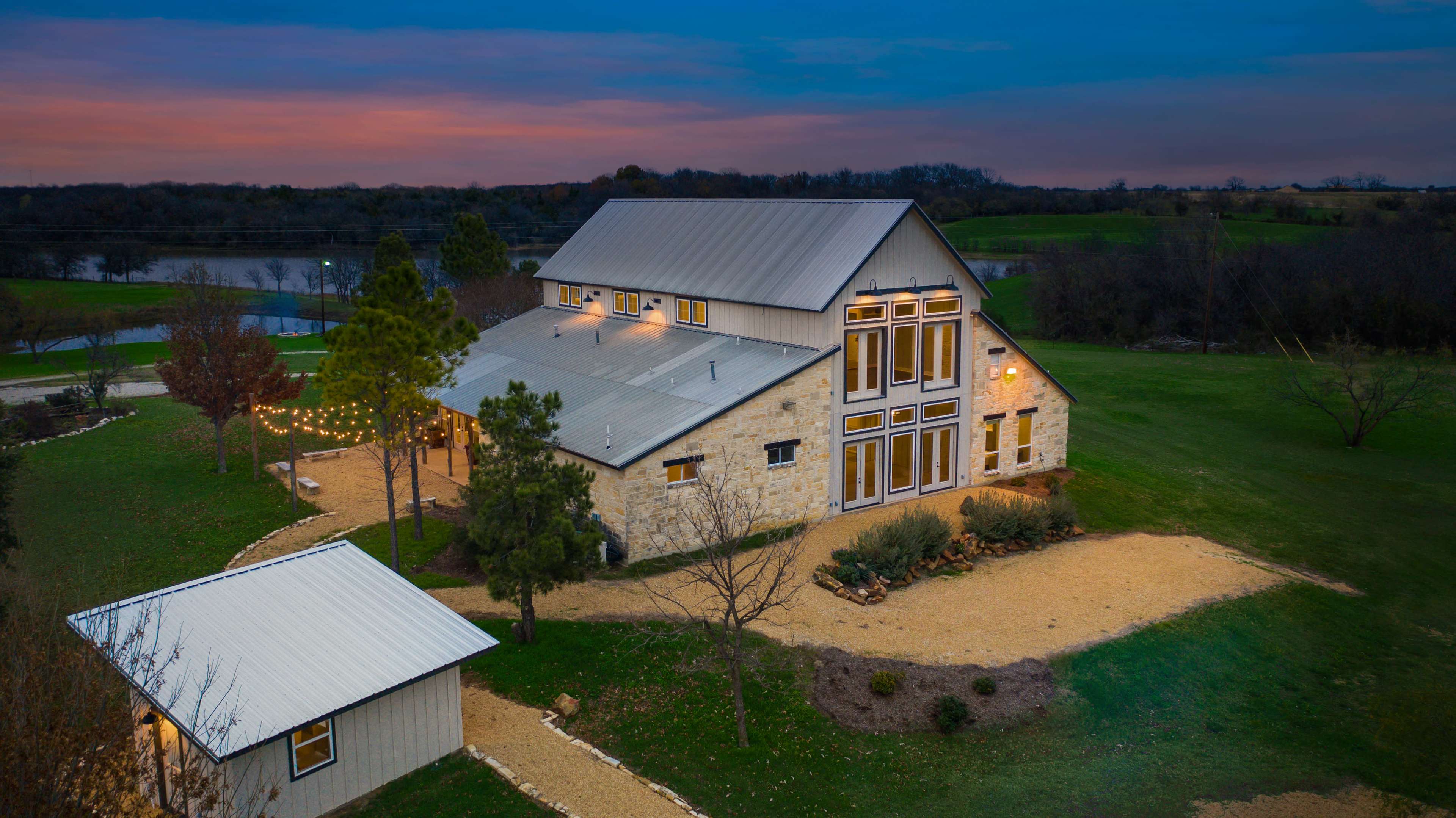The image shows a large, modern barn-style house with a light-colored stone exterior and a metal roof, set in a green landscape at dusk, with warm lights illuminating the exterior.