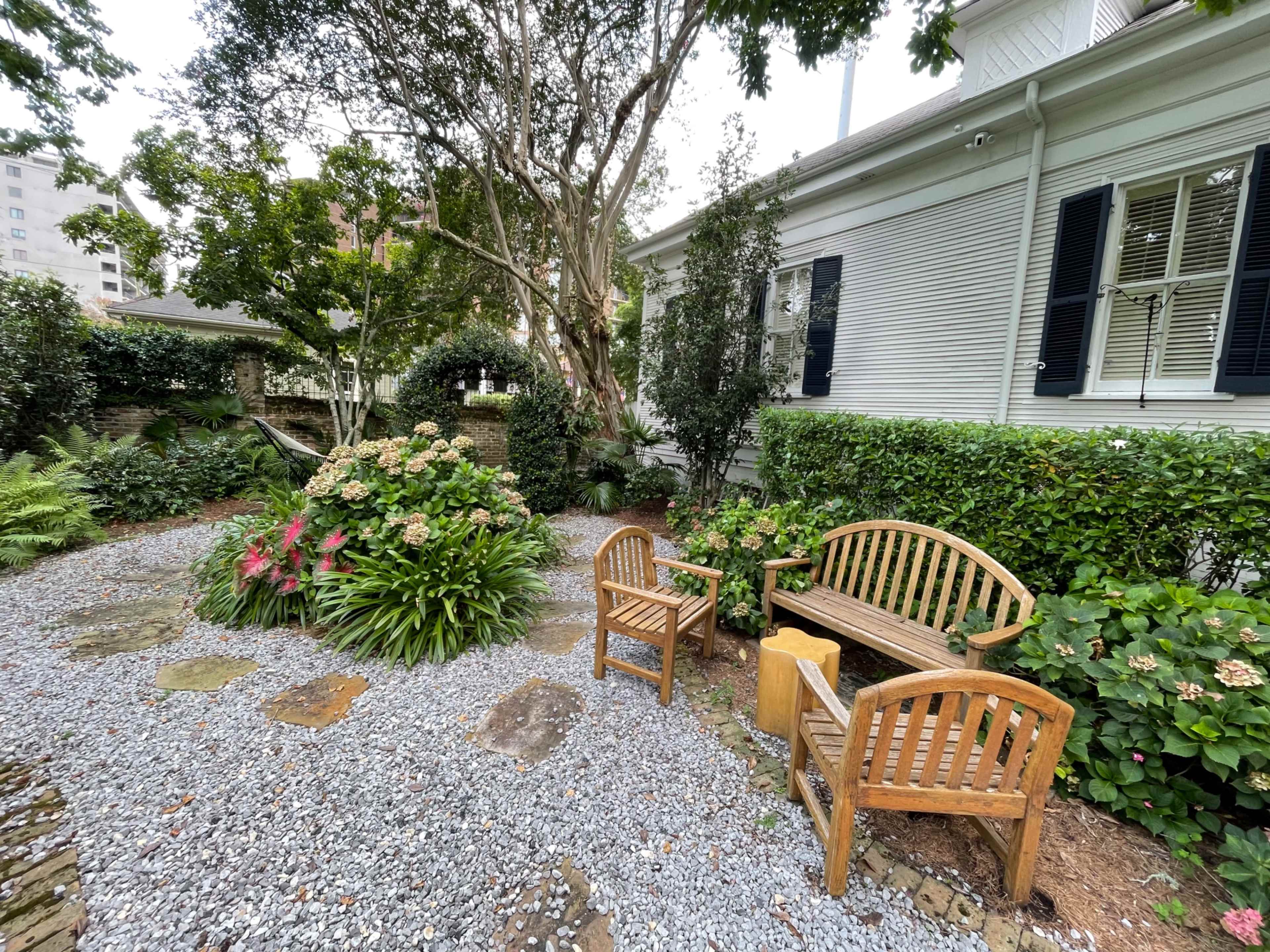 A serene garden area features wooden benches surrounded by lush greenery and flowering plants, with a gravel path leading through the space.