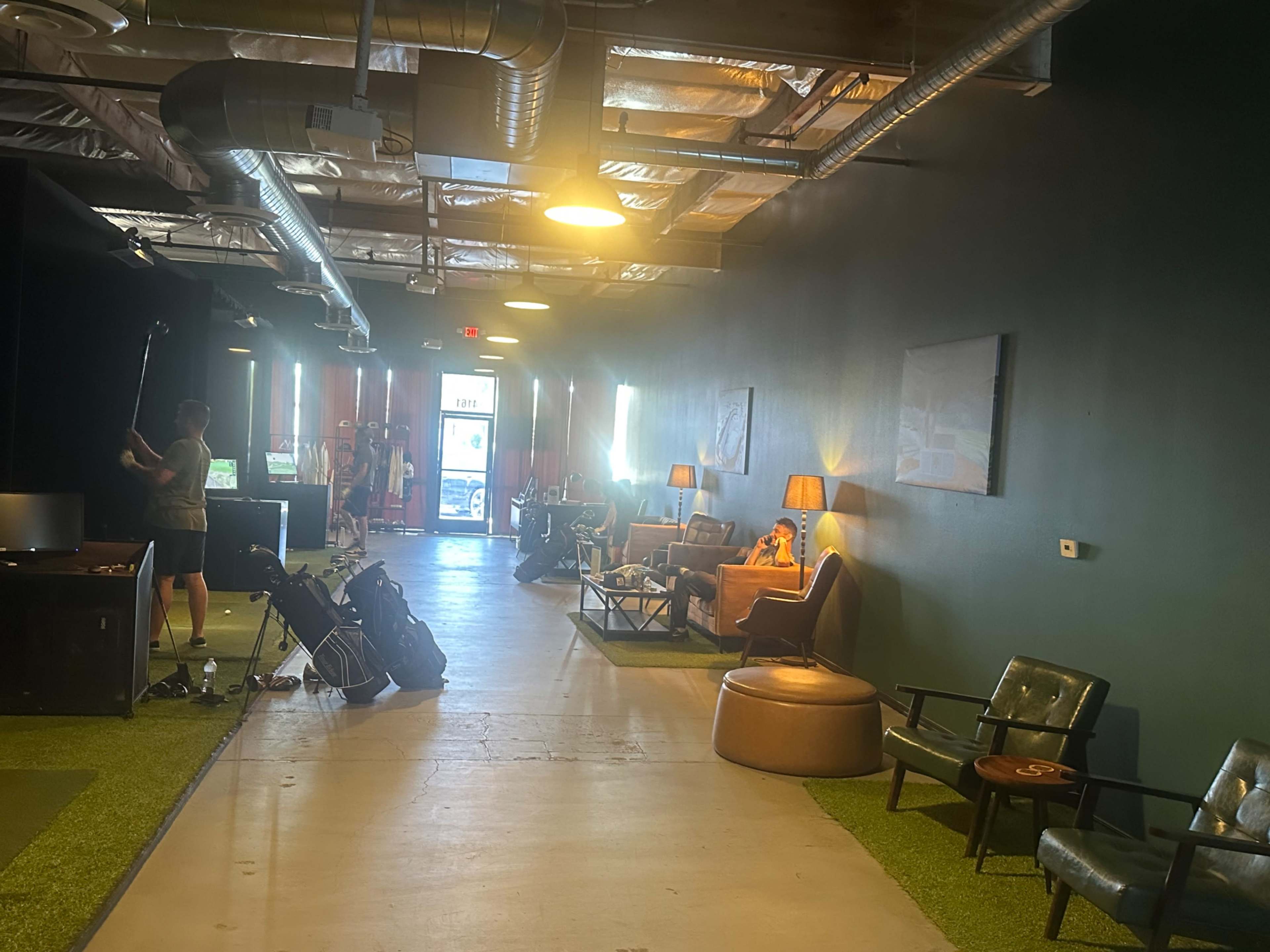 The image shows a spacious hallway with green carpet, multiple seating areas, and individuals engaging in various activities, including golf practice.