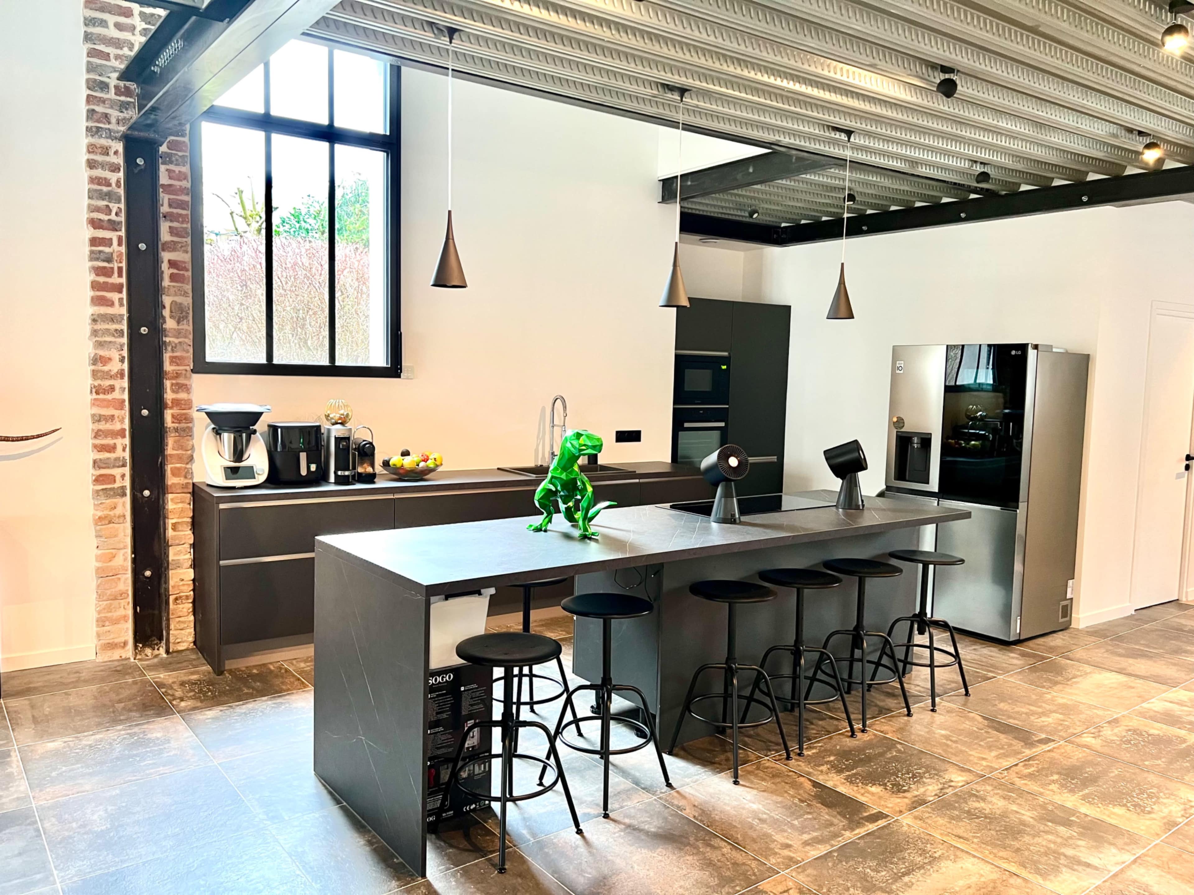 The image shows a modern kitchen with a long dark countertop, bar stools, and appliances including a coffee maker and refrigerator, all set against brick and metal accents.