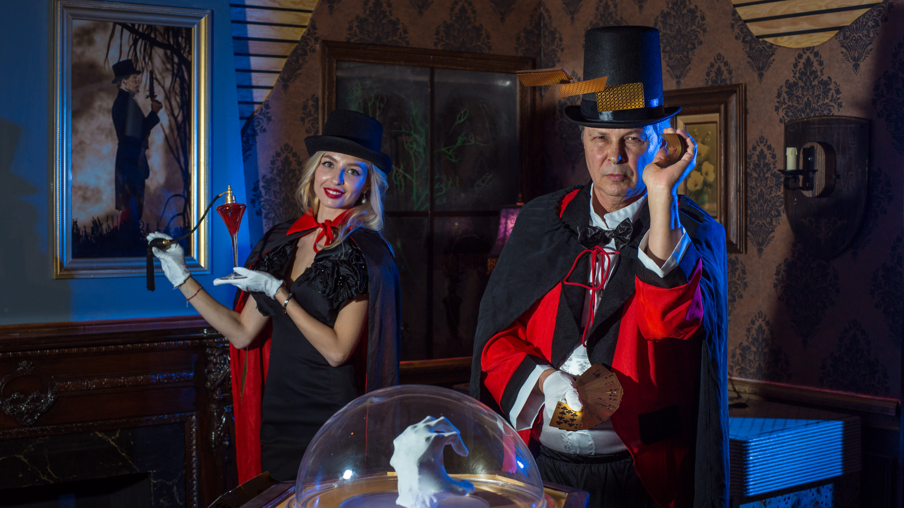 A magician and assistant perform on stage, surrounded by a dramatic set with vintage decor and a glass dome displaying a white rabbit.
