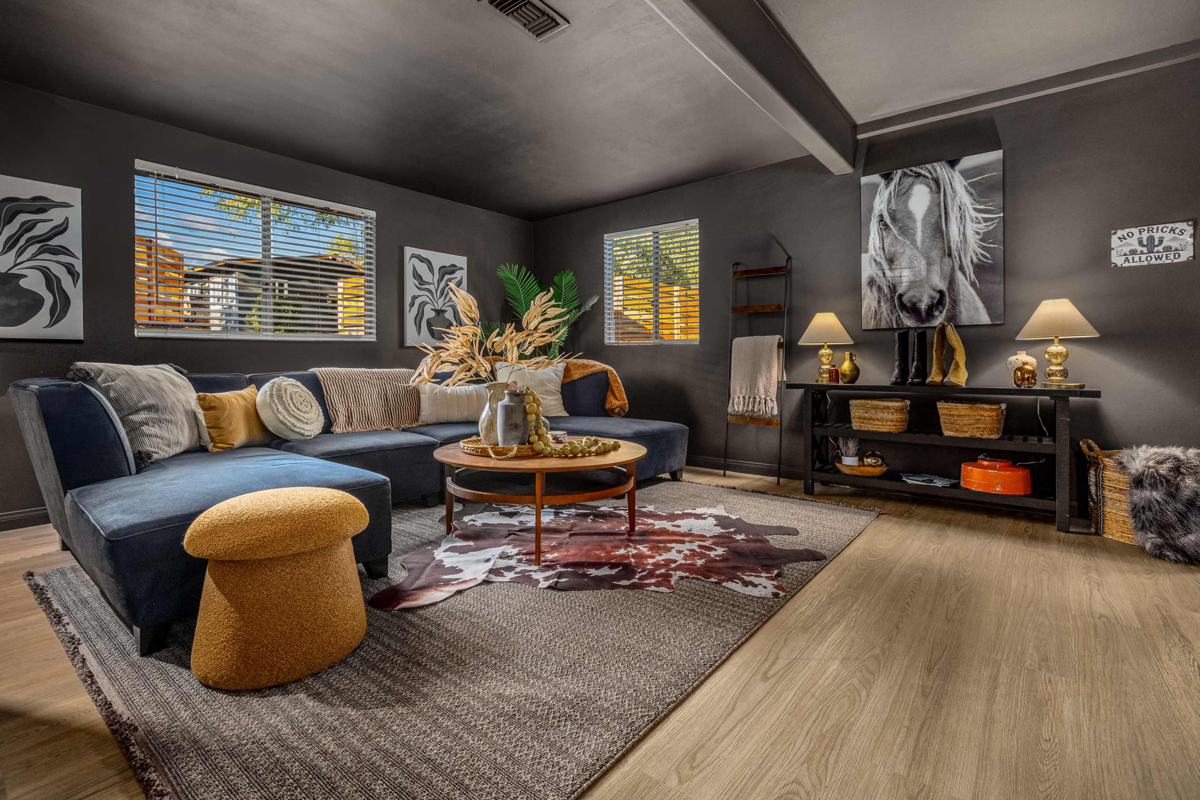 The image shows a modern living room featuring a sectional sofa, a round coffee table, a textured area rug, and wall art, with natural light coming through the windows.