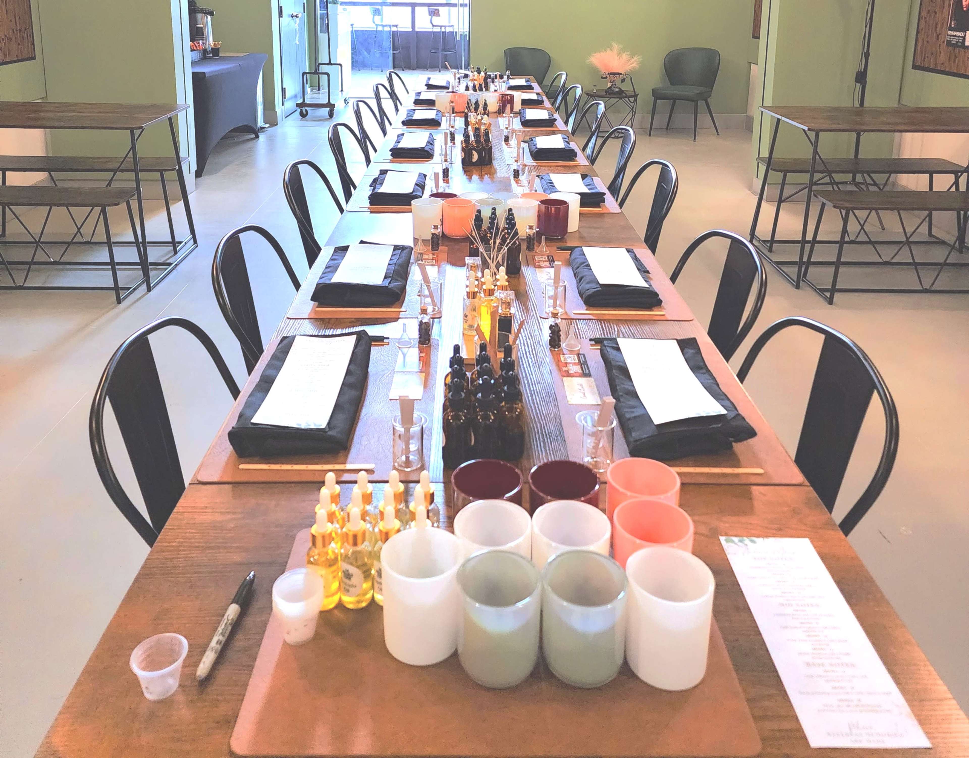 The image shows a long dining table set with black napkins, menus, various bottles, and small containers, arranged in a spacious, well-lit room.