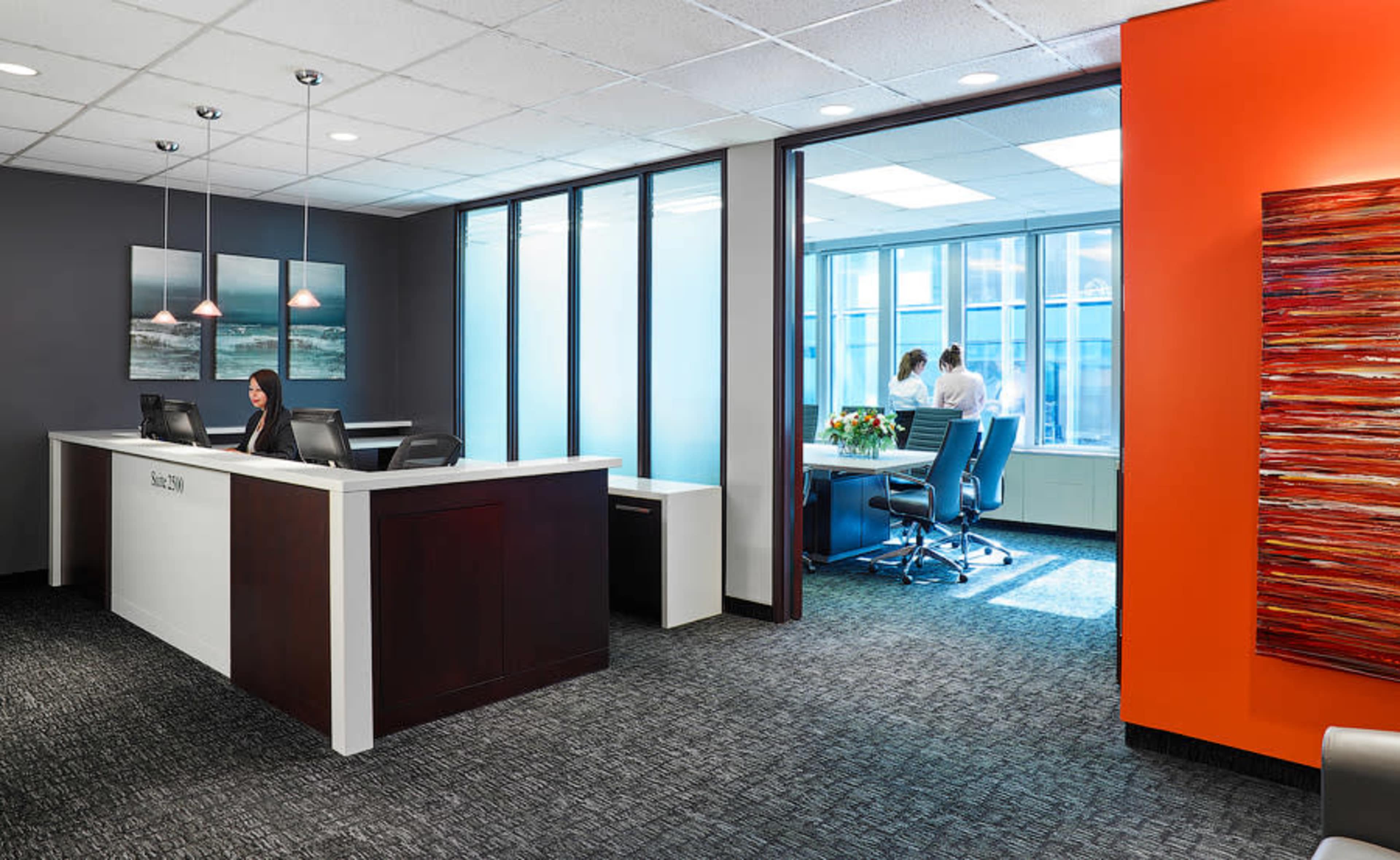 The image shows a modern office reception area with a front desk and a glass partition leading to a meeting room where two people are standing by a window.