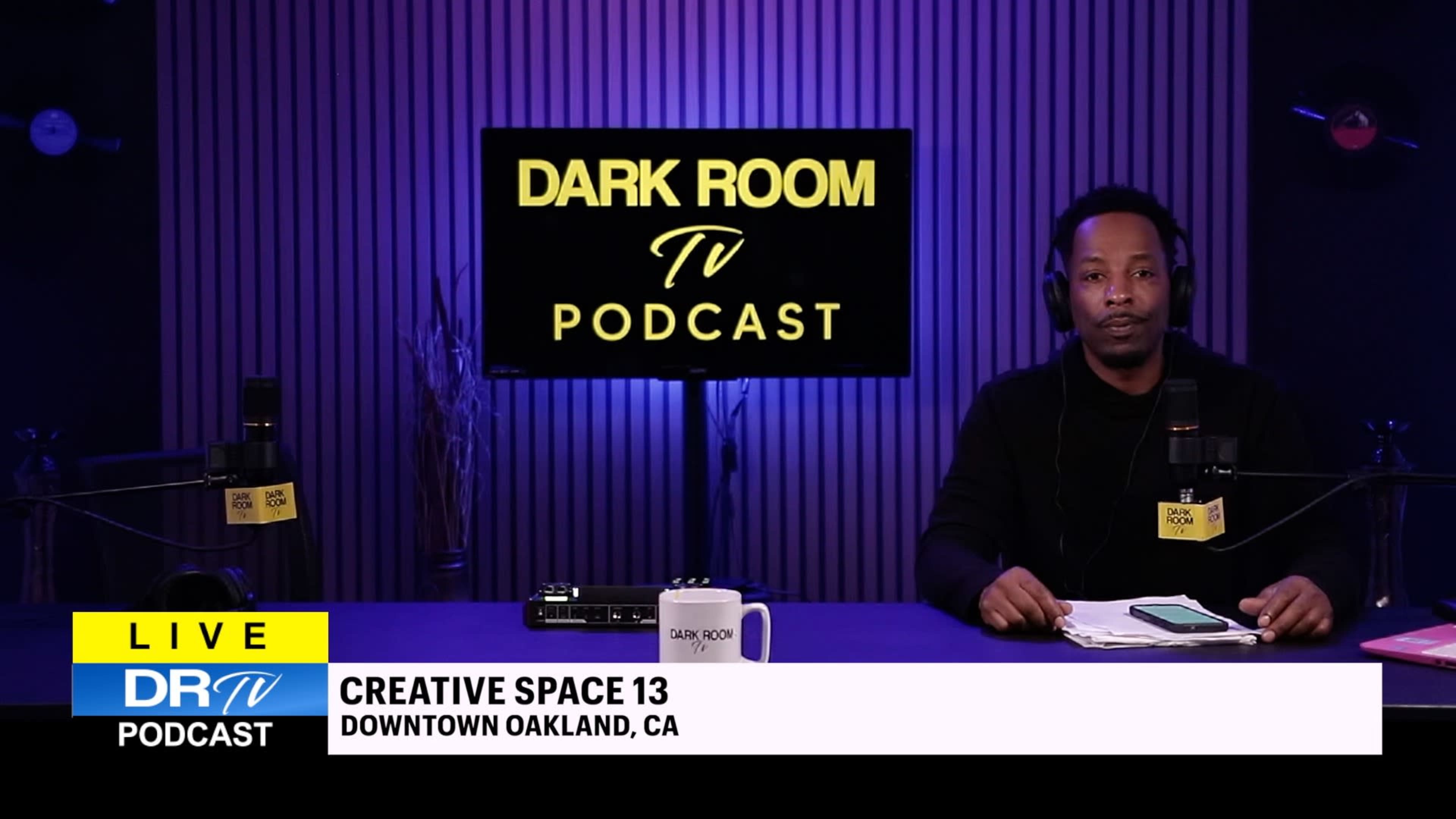 The image shows a man sitting at a table with a microphone and notes, in front of a screen displaying the title "DARK ROOM TV PODCAST" in a purple-lit studio.