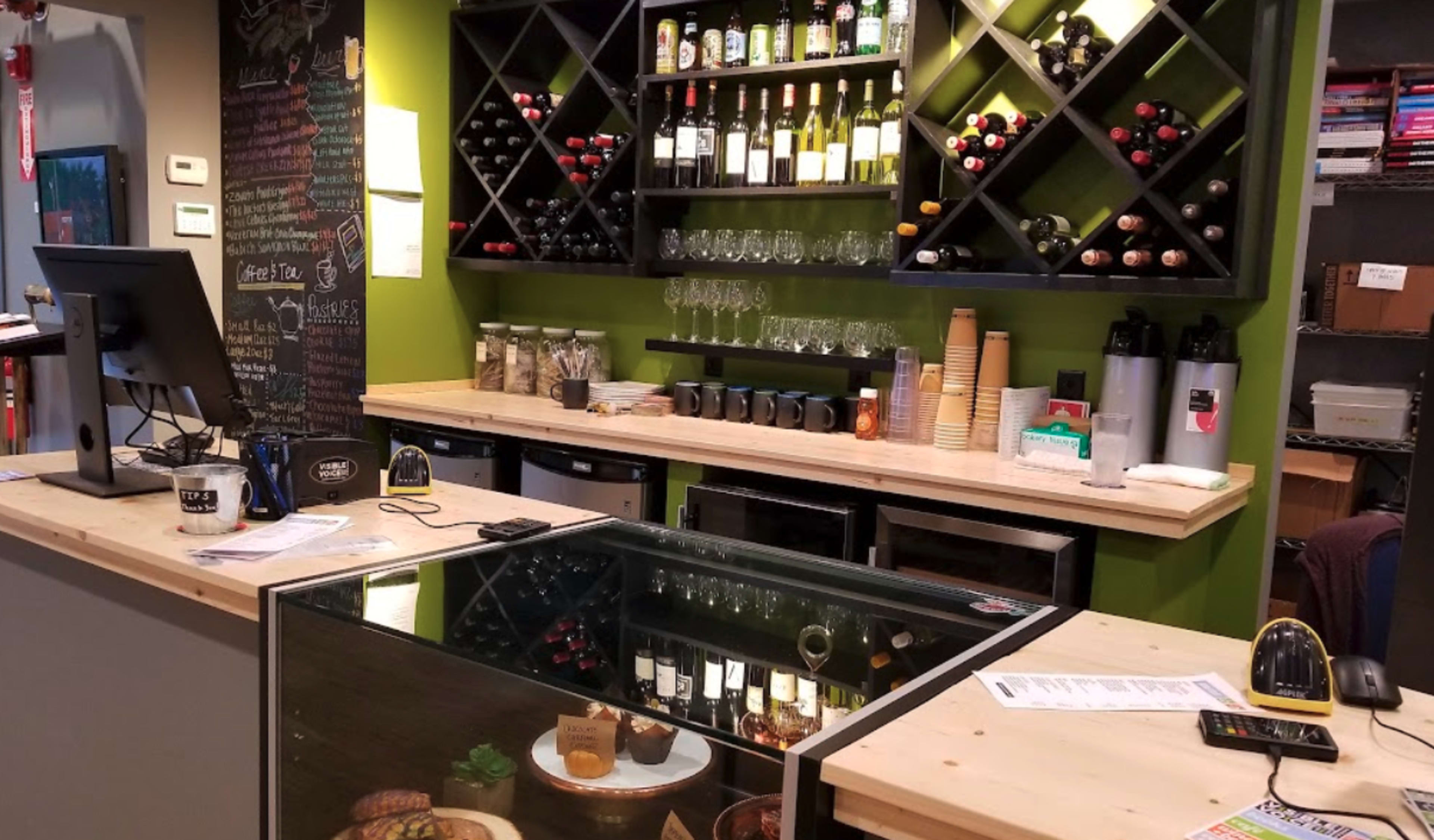 A café counter with a variety of wine bottles on a wall shelf, glassware, a cash register, and a display case showcasing food items.