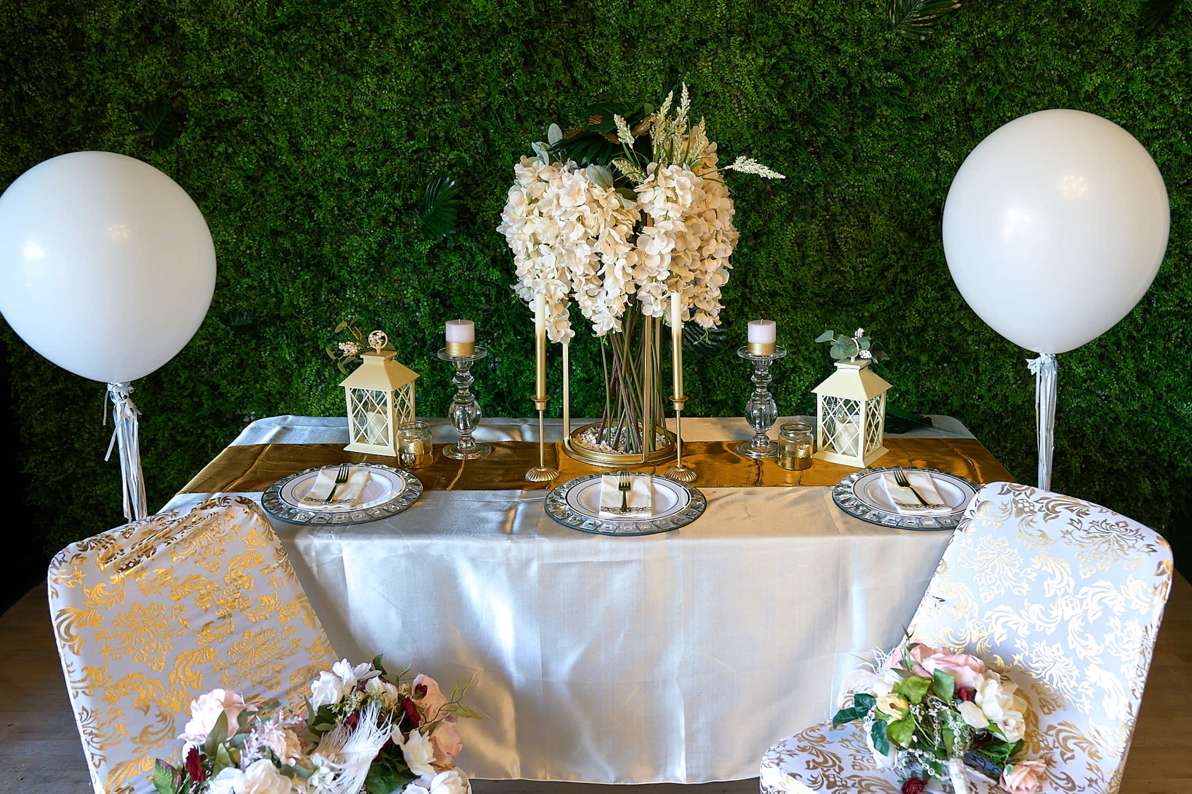 A dining table is elegantly set with floral centerpieces, candles, and decorative lanterns, surrounded by two chairs adorned with floral arrangements and large balloons on either side.