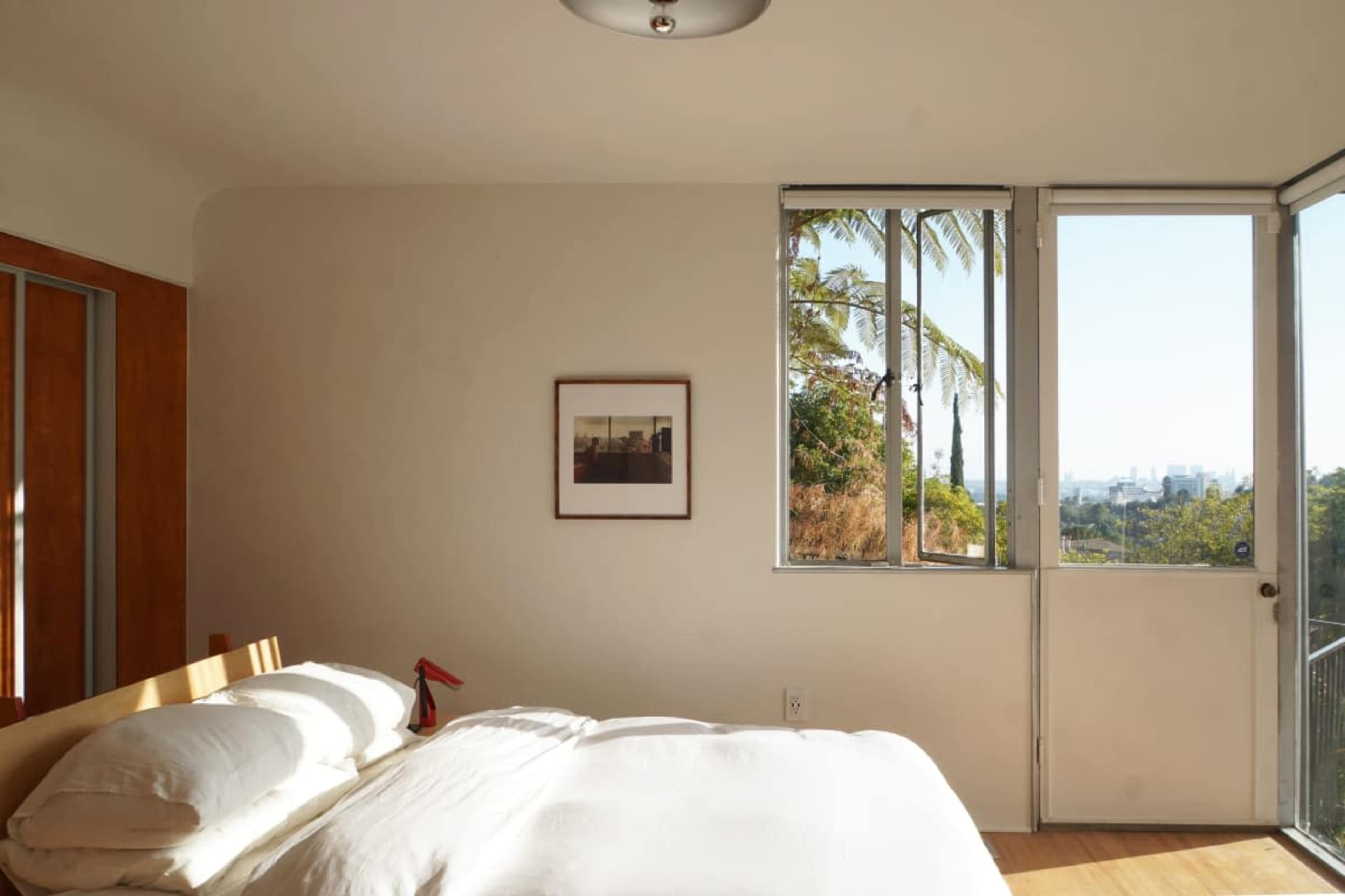 A bright bedroom features a bed with white linens, a framed picture on the wall, and large windows that open to a view of greenery and the city beyond.