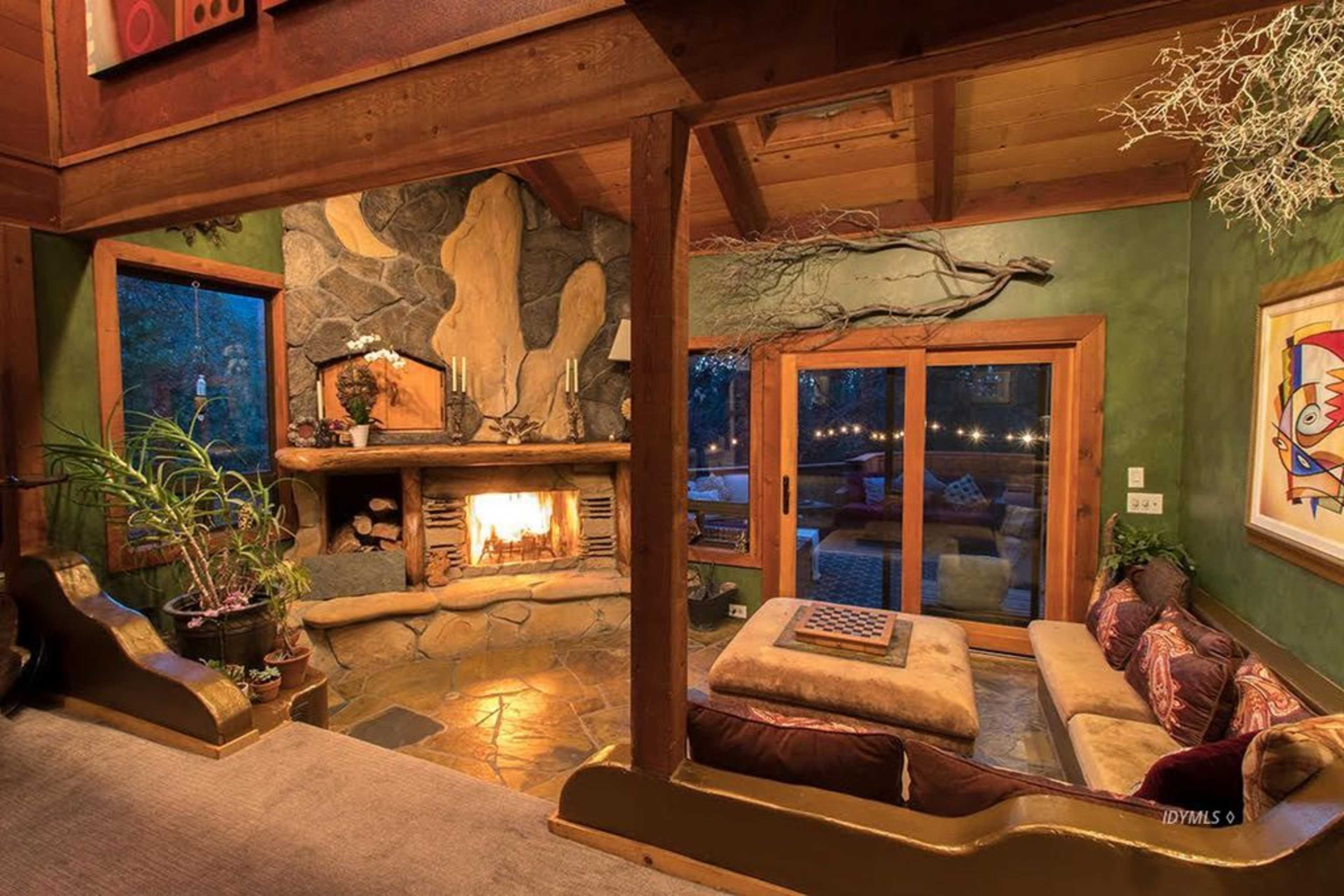 The image shows a cozy living room with a stone fireplace, wooden beams, and large windows overlooking an outdoor area.