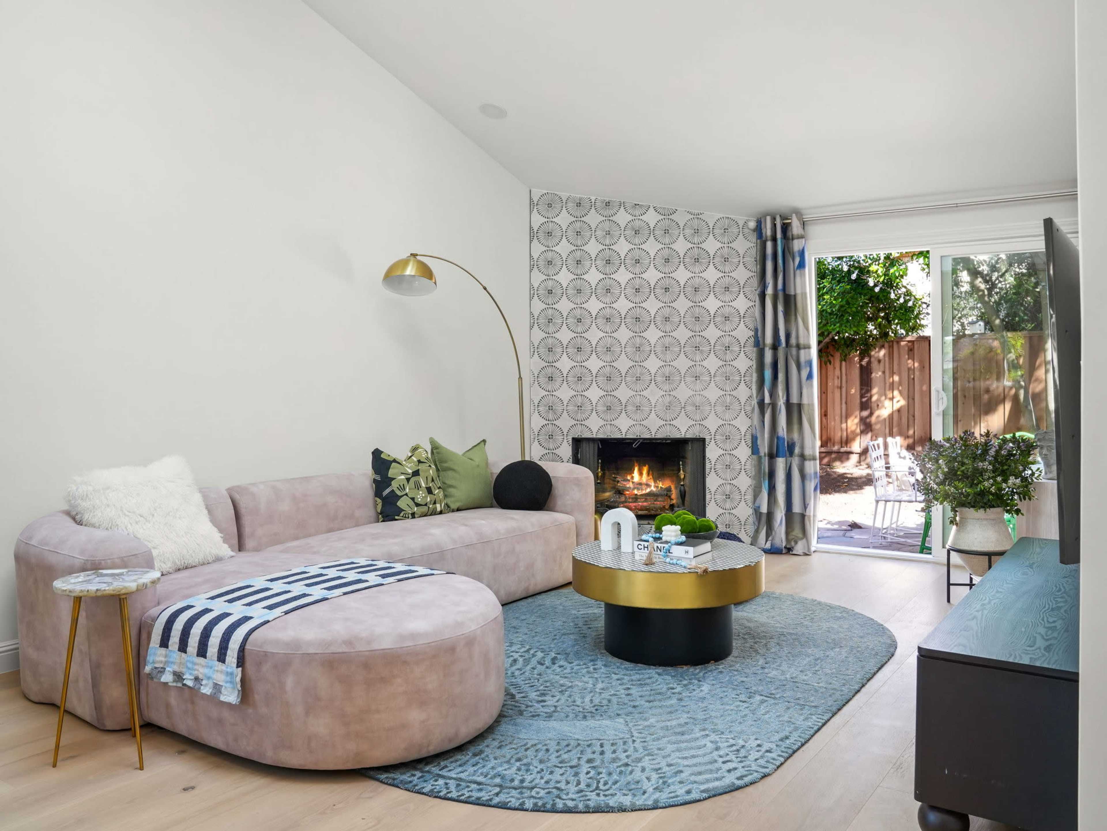 A modern living room features a curved sofa, a round coffee table, and a fireplace, with large windows opening to an outdoor space.
