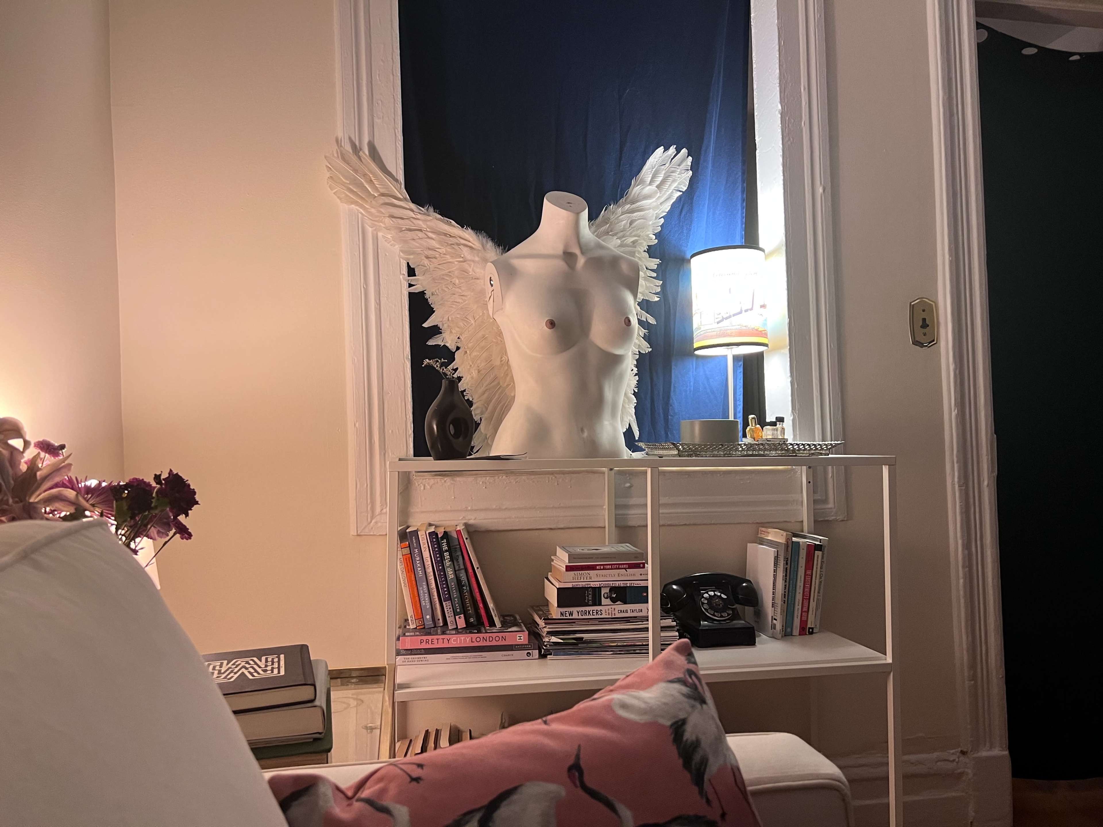 A winged mannequin placed near a dimly lit bookshelf and a lamp with a blue curtain in the background.