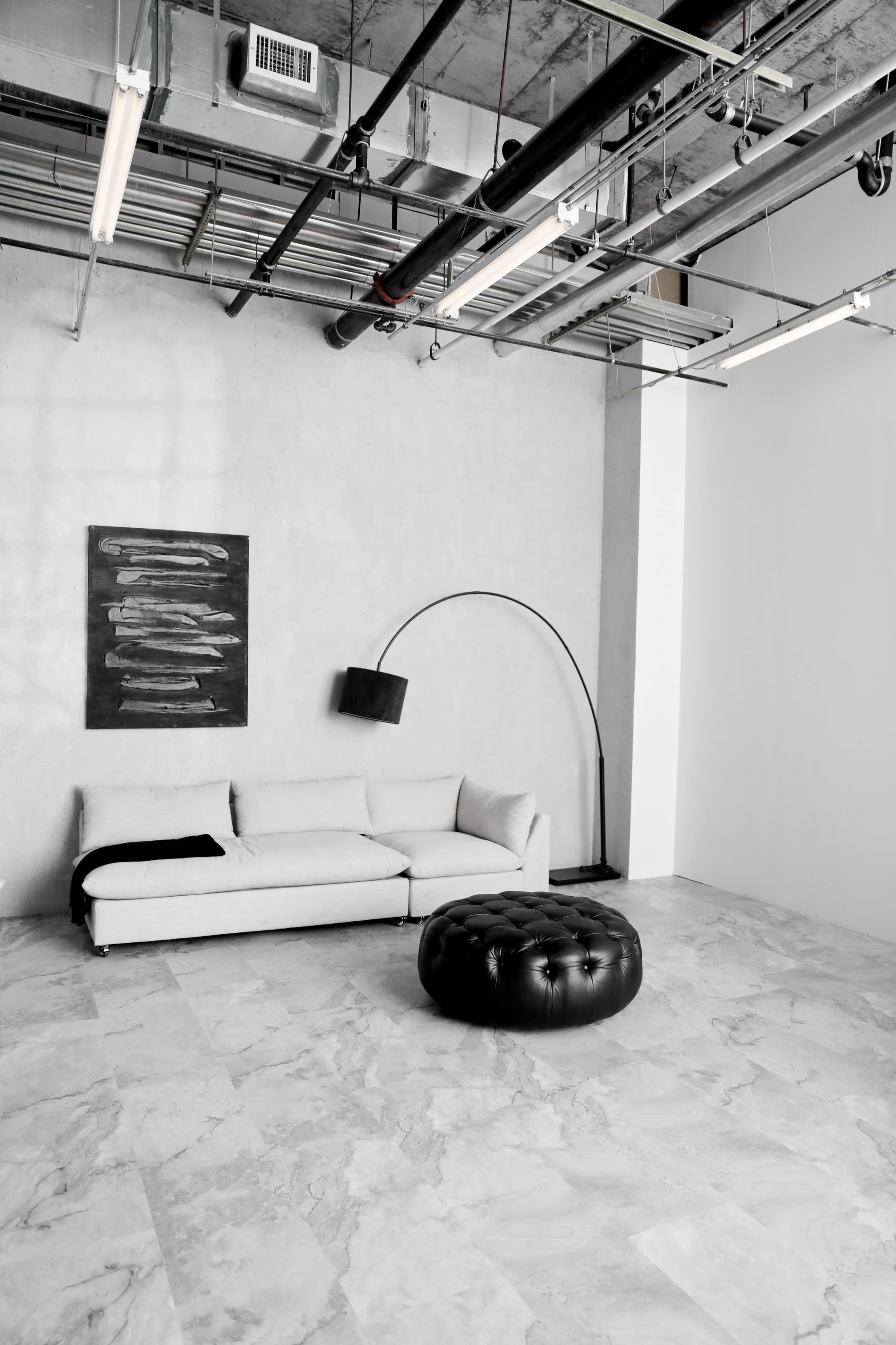 A minimalist living space with a gray wall, a white sofa, a black floor lamp, and a black tufted ottoman on a marble floor.