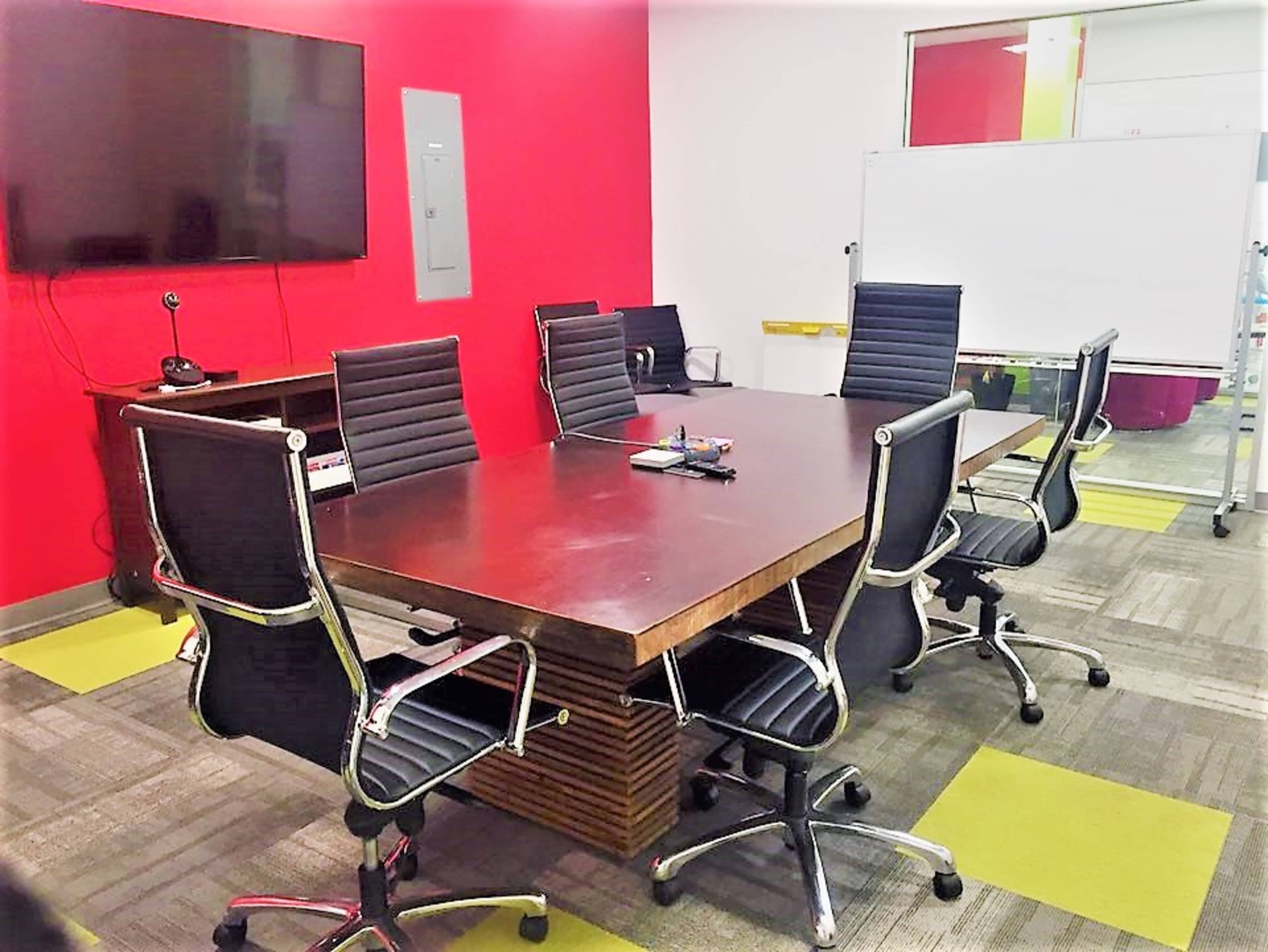 A modern conference room features a large wooden table surrounded by black rolling chairs, with a television mounted on a red wall and a whiteboard nearby.