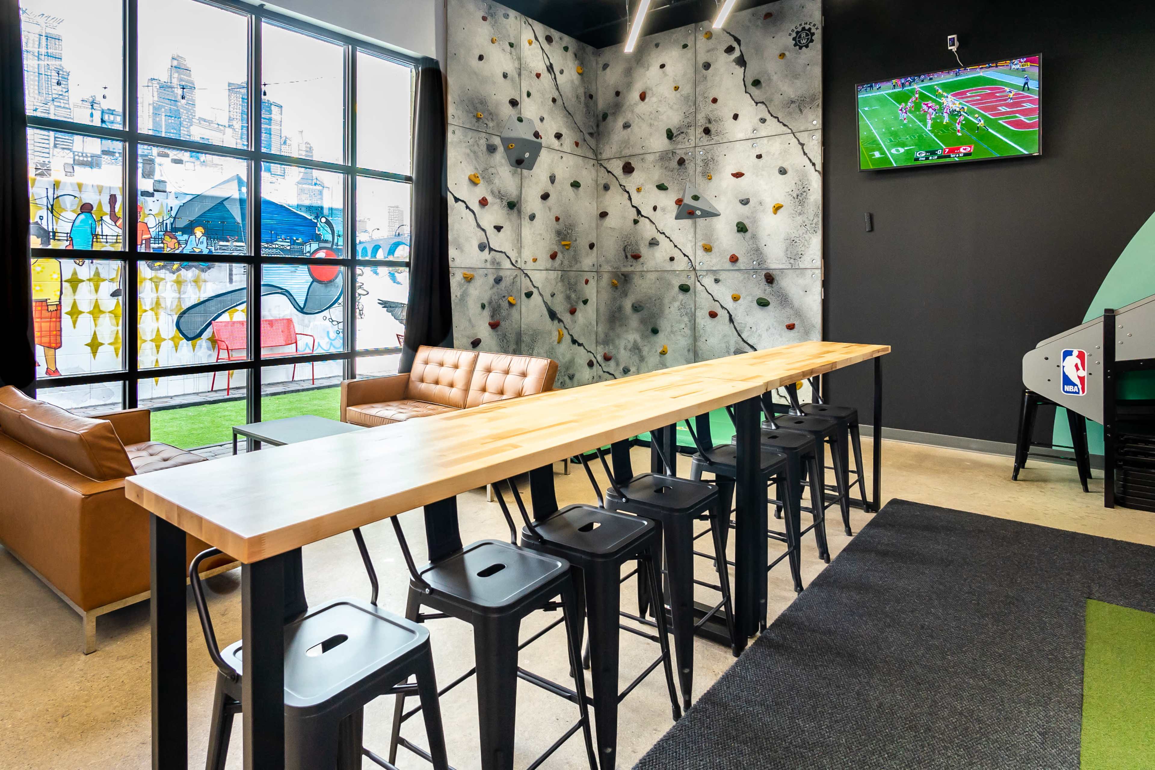 The image features a modern recreational space with a wooden bar, black stools, a climbing wall, a television displaying a sports game, and large windows showcasing urban murals.