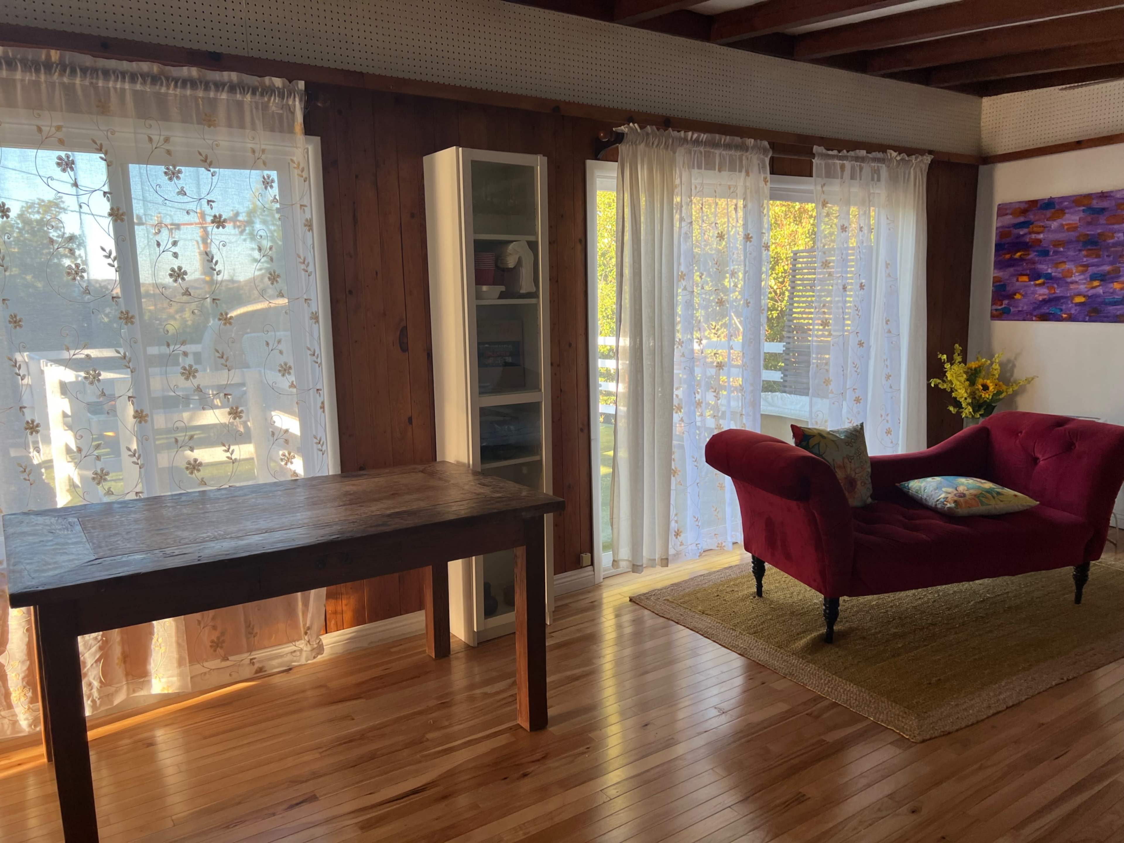 The image shows a room with wooden flooring, a wooden table, a red upholstered sofa, sheer curtains, and a bookshelf by a window that lets in natural light.