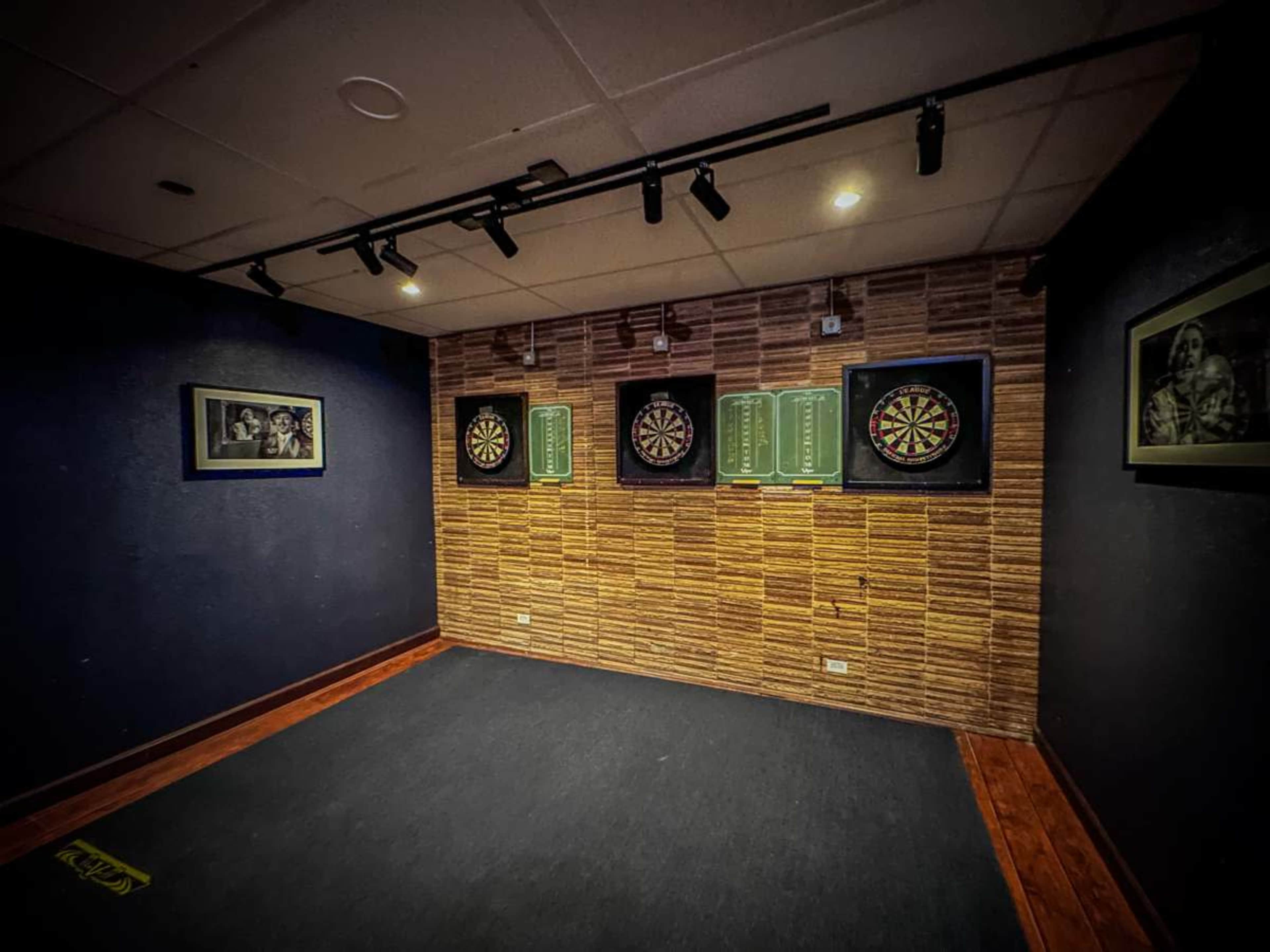 The image shows a dart room with three dartboards mounted on a textured wall, illuminated by track lighting.