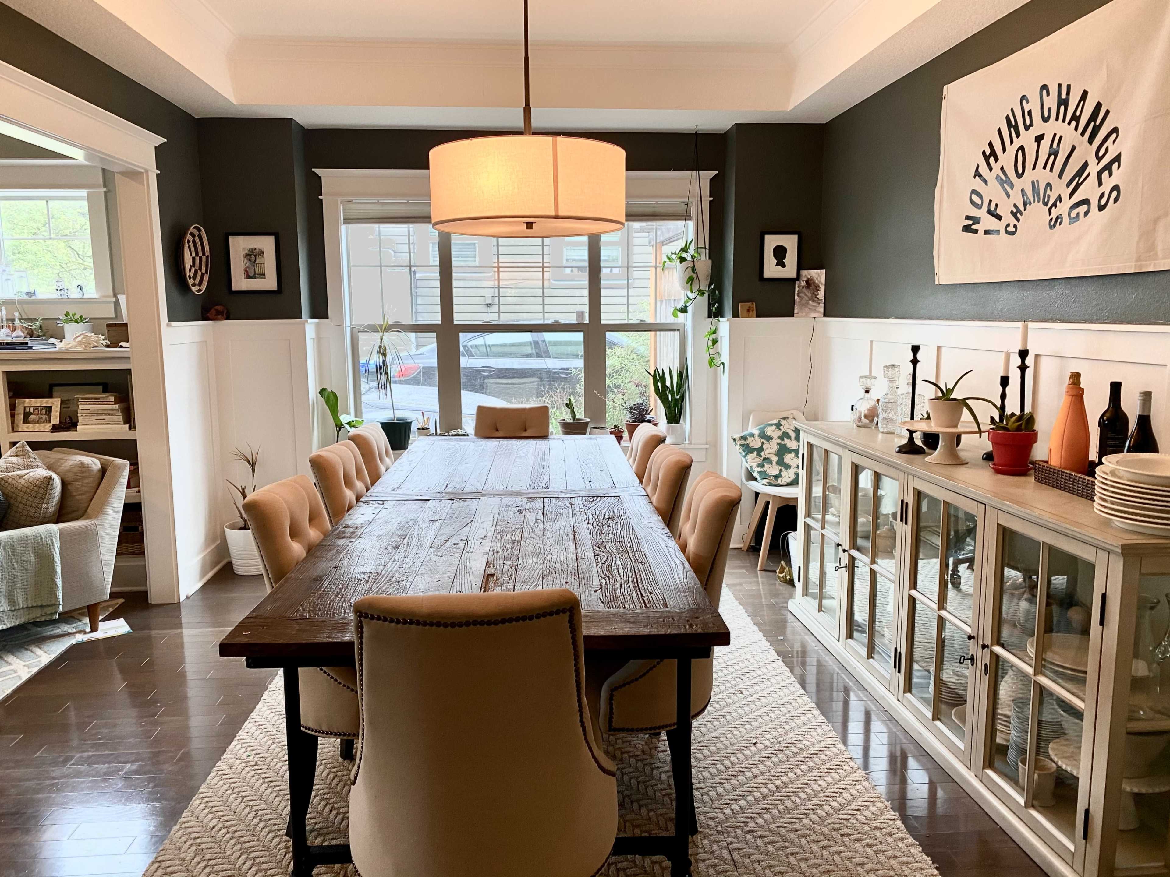 A spacious dining room features a long wooden table surrounded by upholstered chairs, with a large window providing natural light and shelves displaying various decorative items.