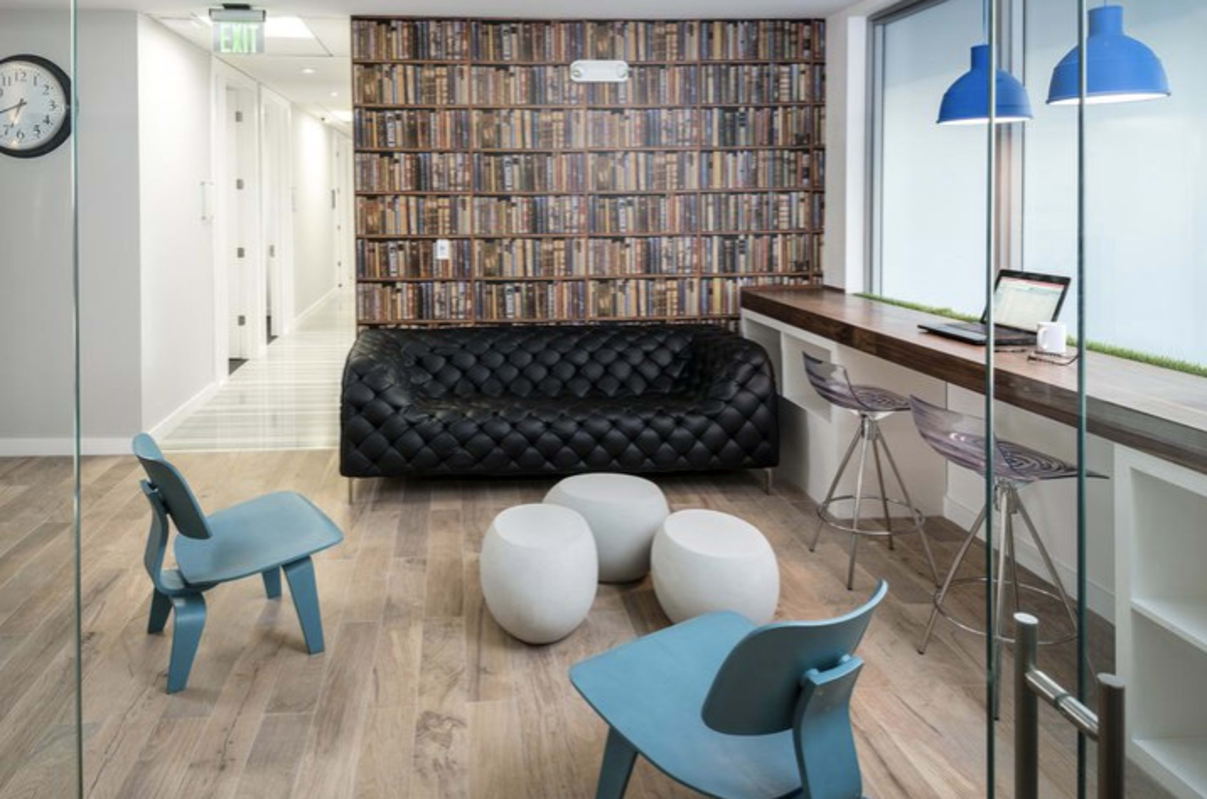 A modern office lounge with a black tufted sofa, two light blue chairs, and a wooden countertop with bar stools, all set against a wall featuring a bookshelf wallpaper.