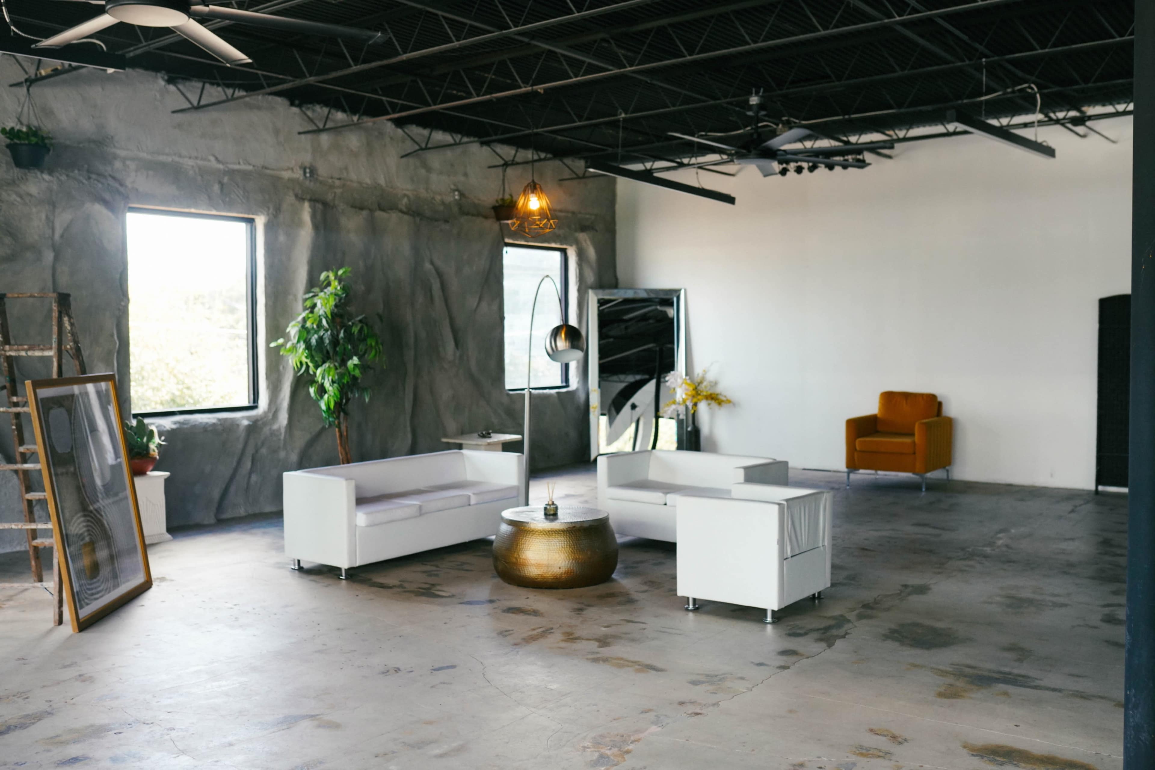 The image shows a spacious, industrial-style room with concrete walls, large windows, and minimal furniture, including white sofas and a golden coffee table.