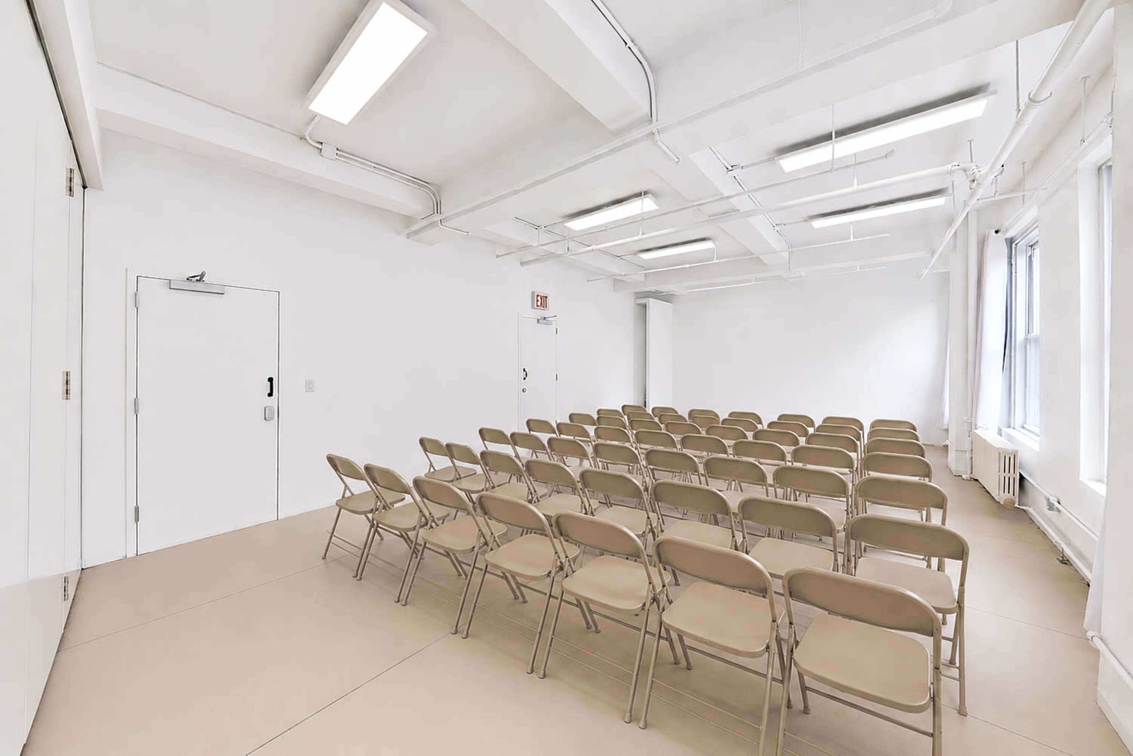 The image shows a minimalist, empty room with rows of beige chairs facing a blank wall, illuminated by overhead lights.