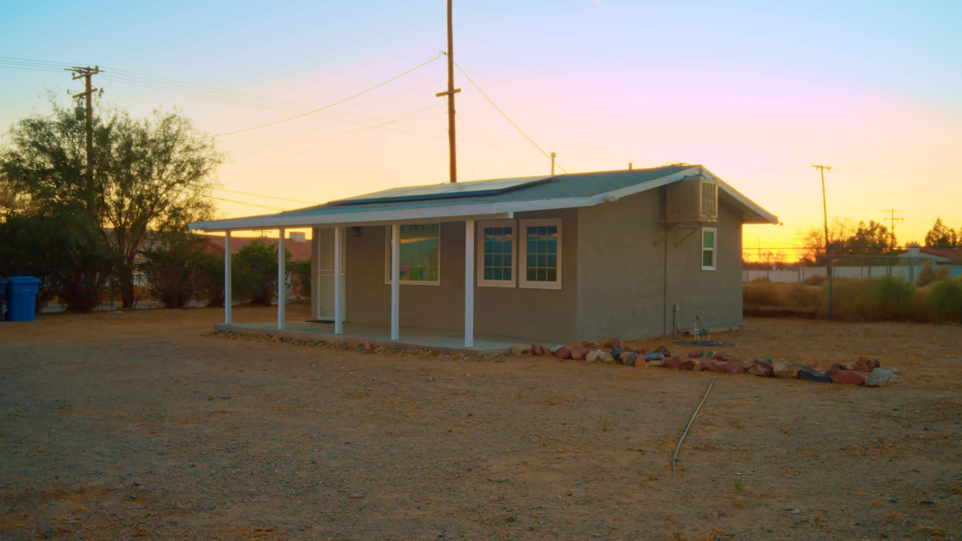 A single-story gray house with a front porch is set on a gravel lot under a colorful sunset sky.