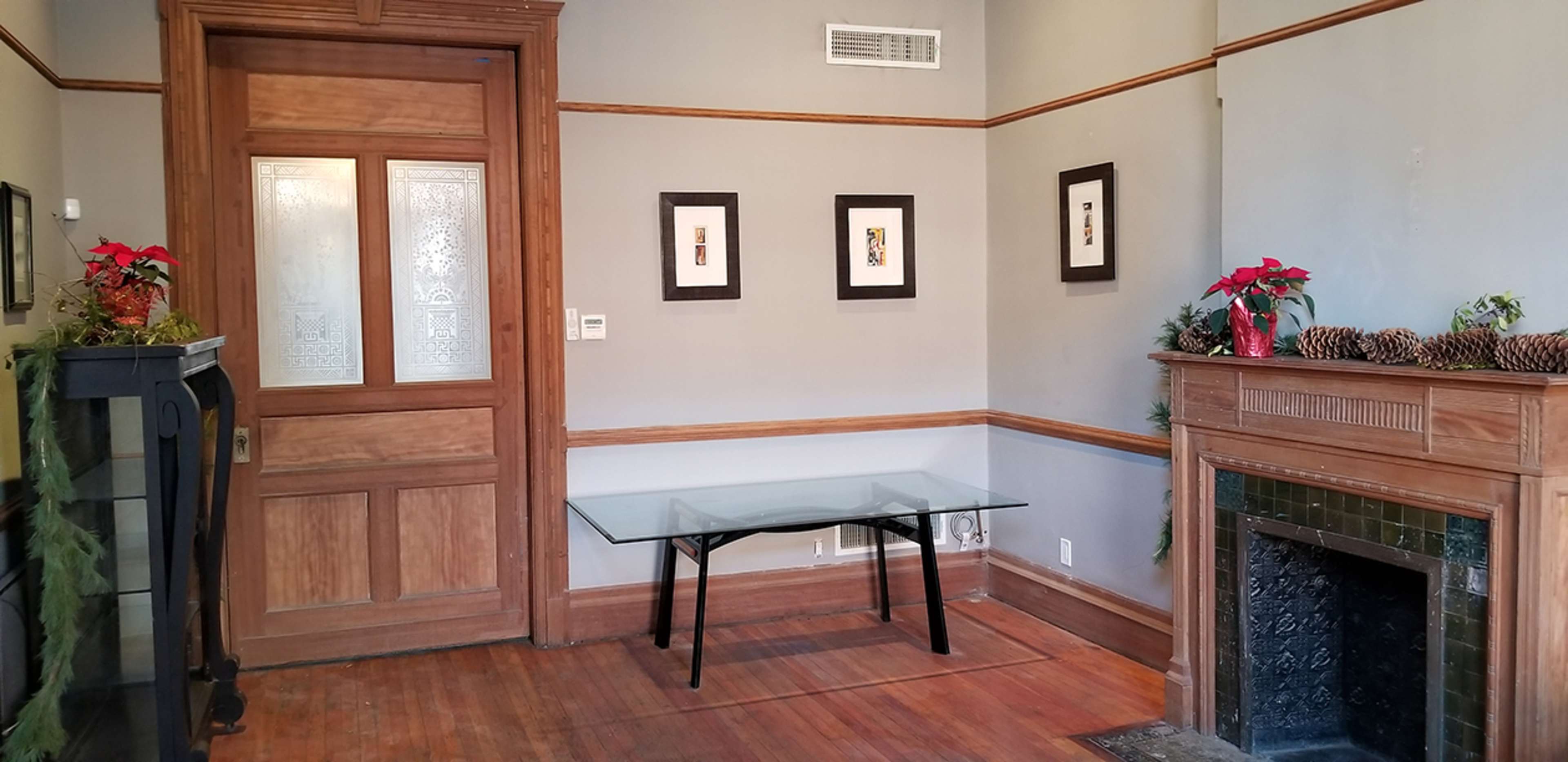 The room features a glass table against a wall with framed artwork, a wooden fireplace with pinecones and plants, and a decorative door.
