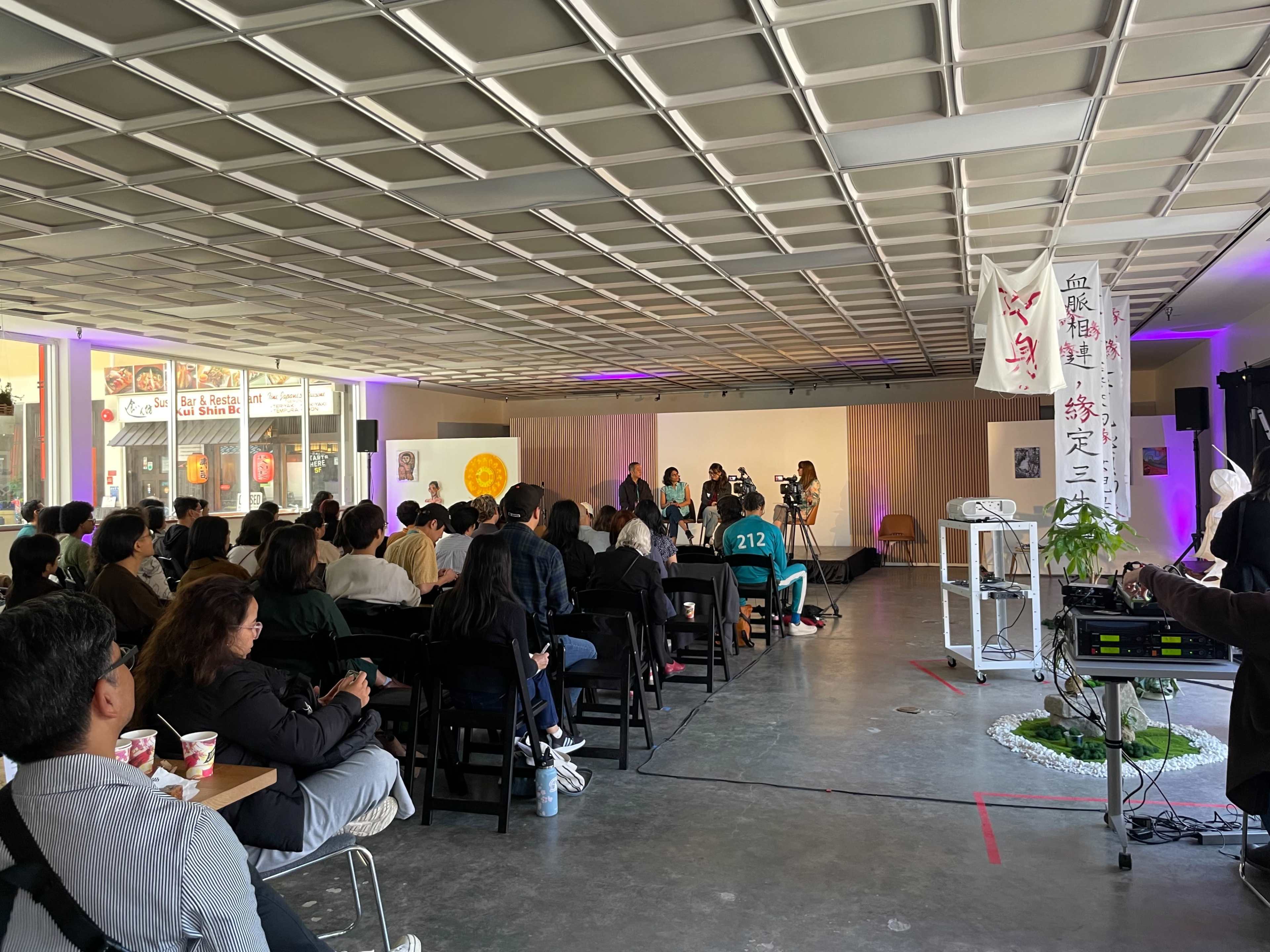 A seated audience watches a panel discussion taking place on a stage in a well-lit indoor space.