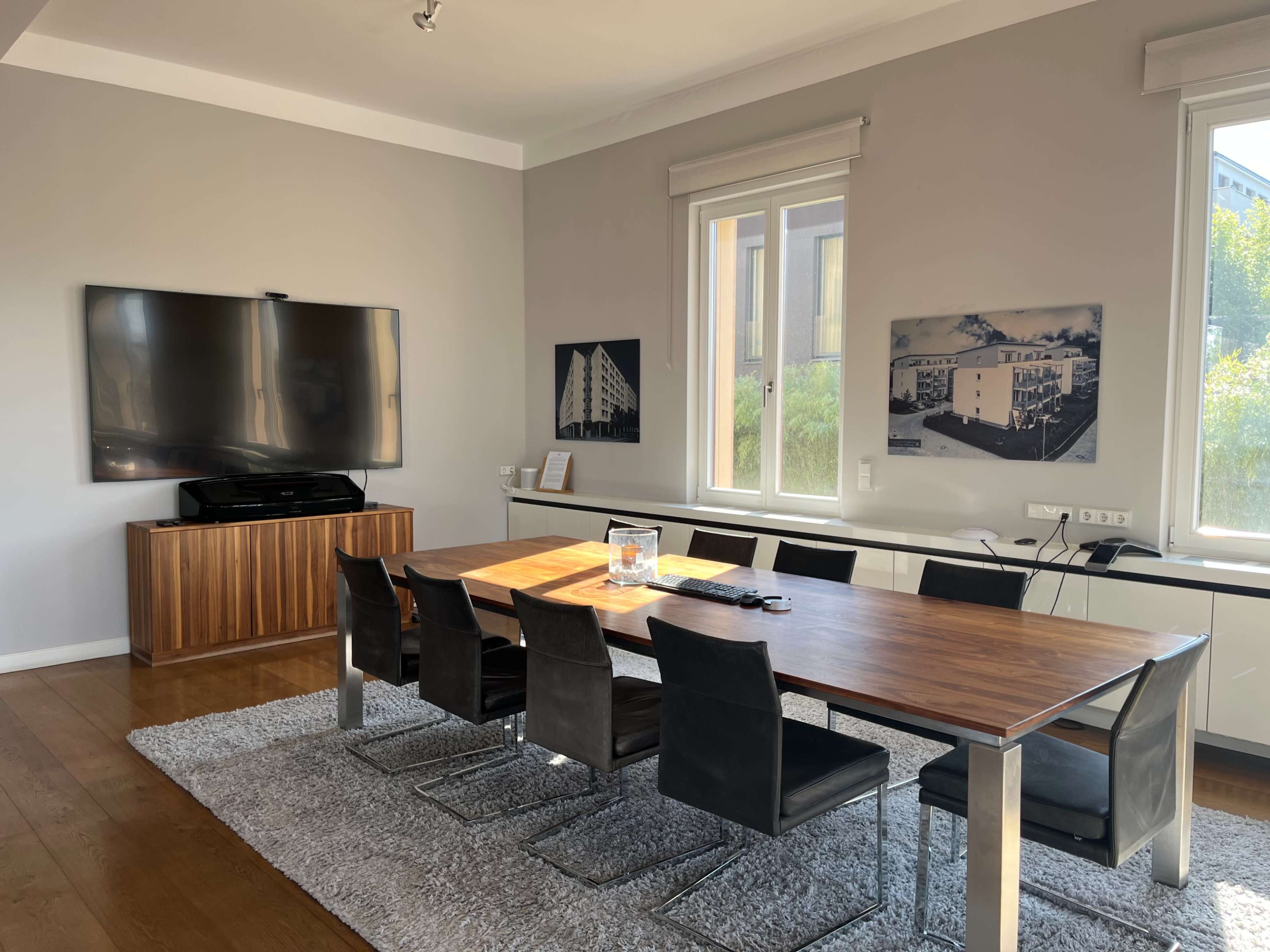 The image shows a modern dining area featuring a long wooden table surrounded by black chairs, with a large television mounted on the wall and artwork displayed beside windows that allow natural light into the room.