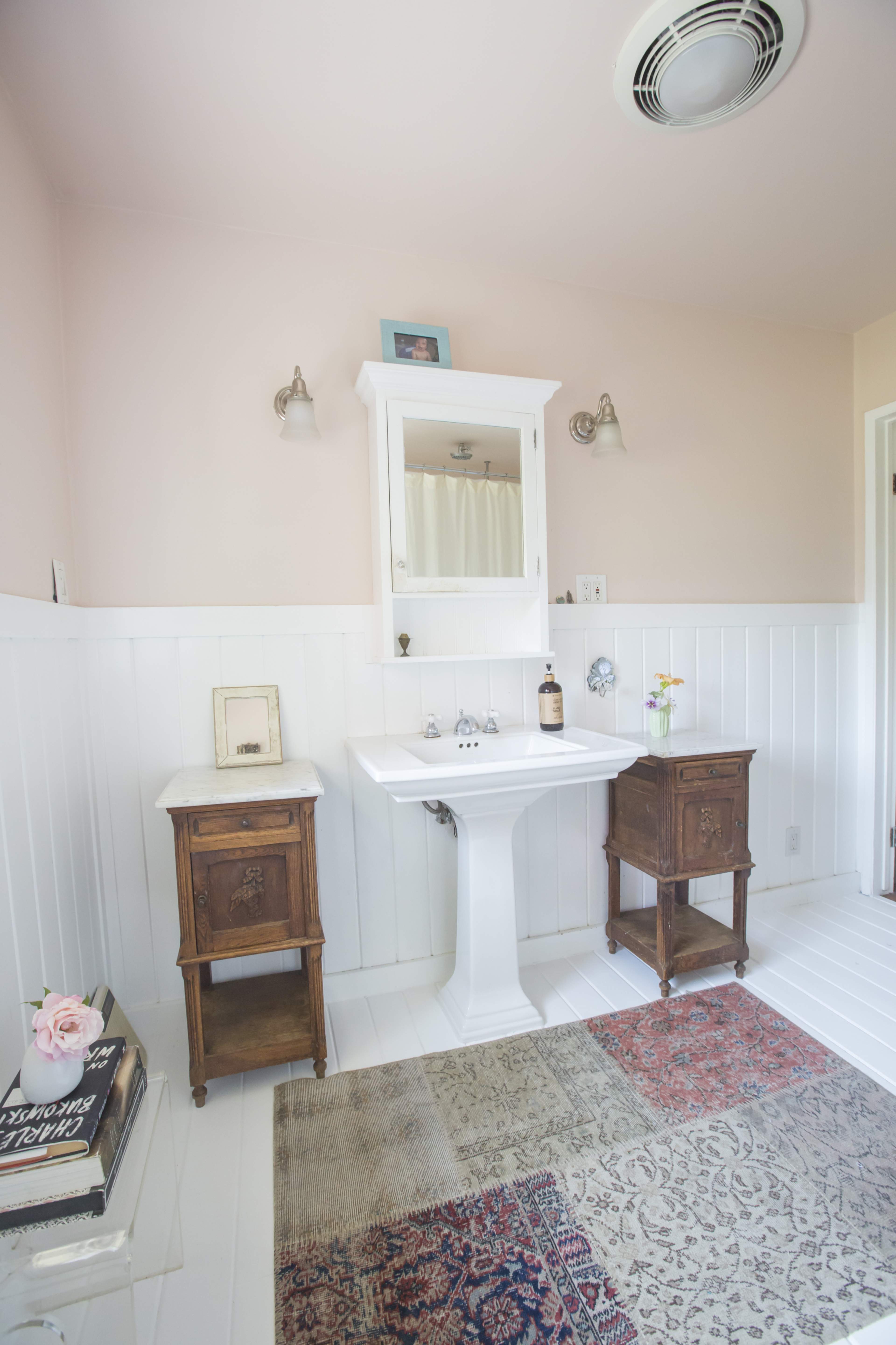 A bright bathroom features a white pedestal sink, two wooden nightstands, and a patterned rug.