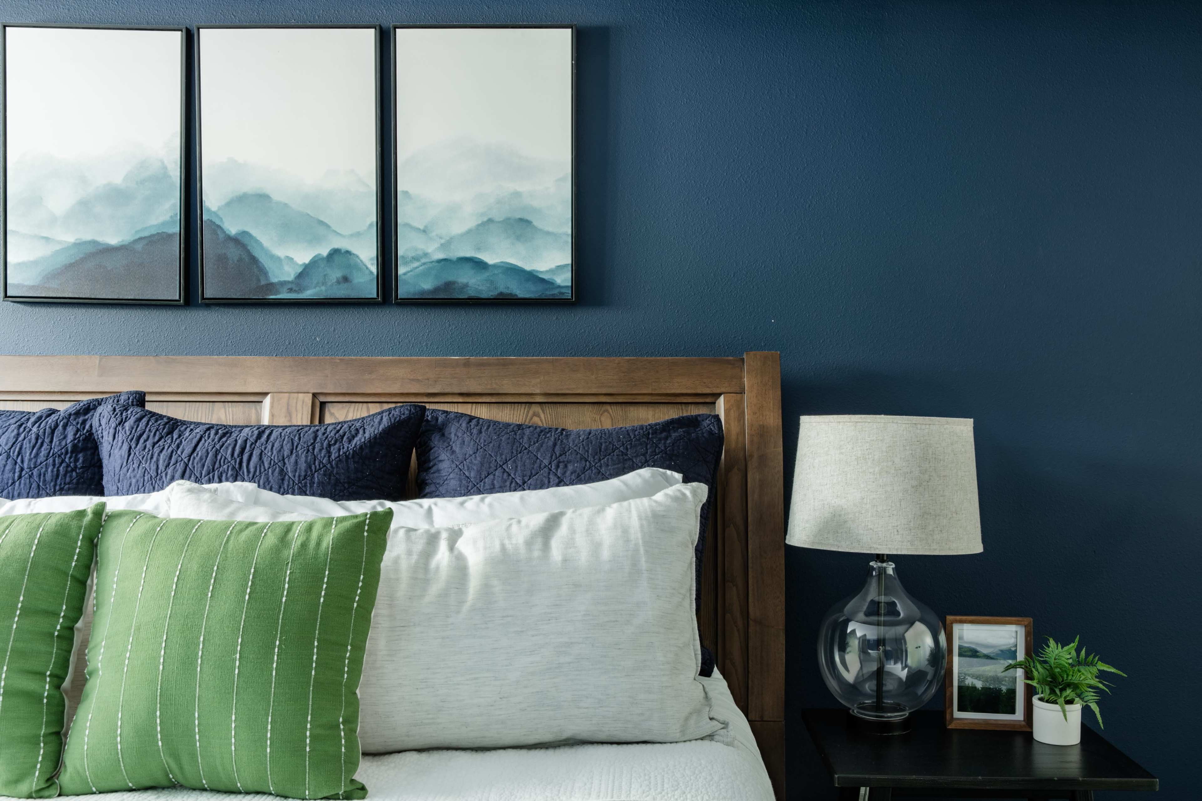 The image shows a modern bedroom with a wooden bed frame, decorative pillows in green and navy, and three landscape paintings on a dark blue wall.