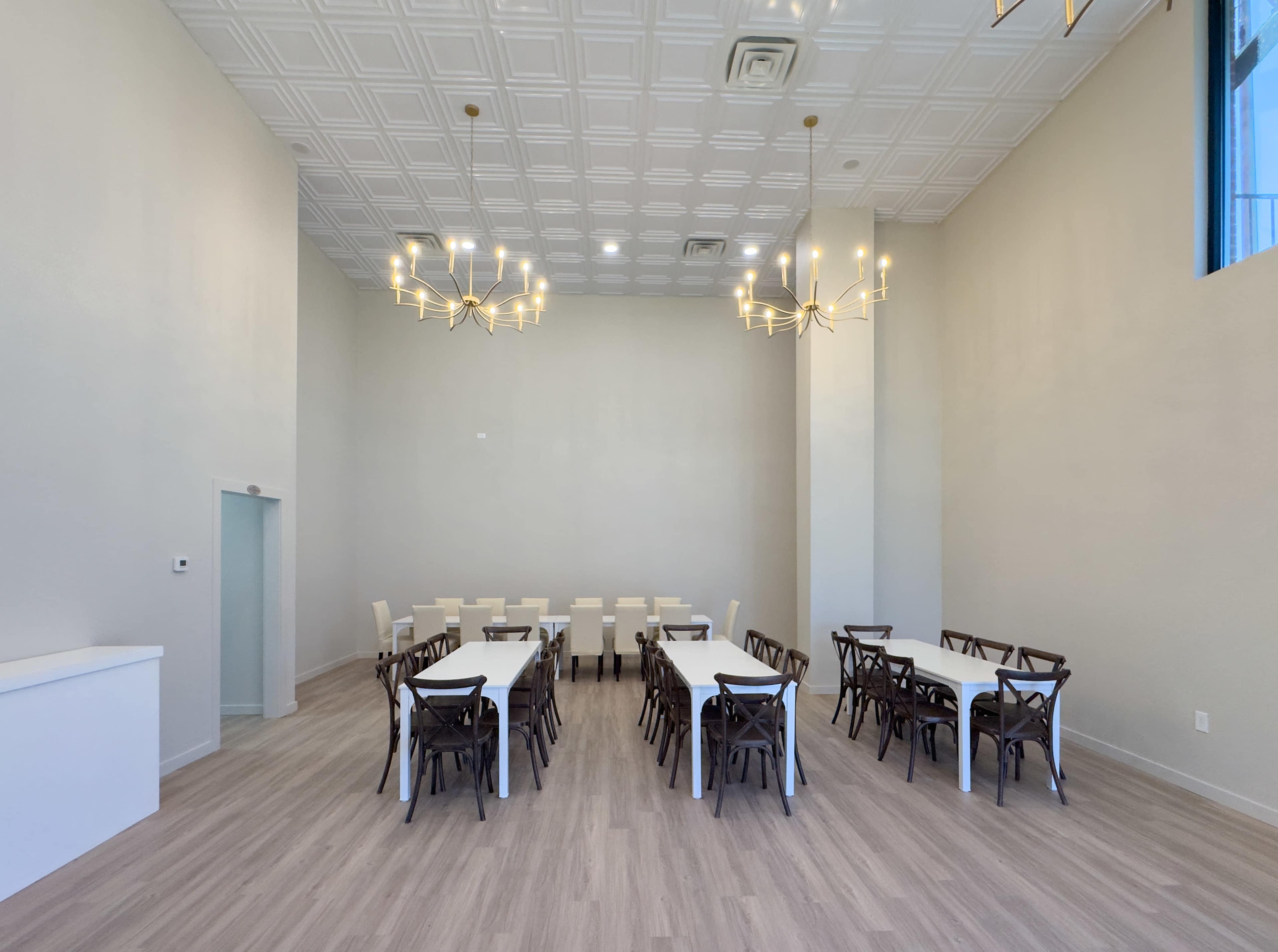 The image shows a spacious, empty room with white walls, two large chandeliers, and rows of tables and chairs arranged for a gathering.
