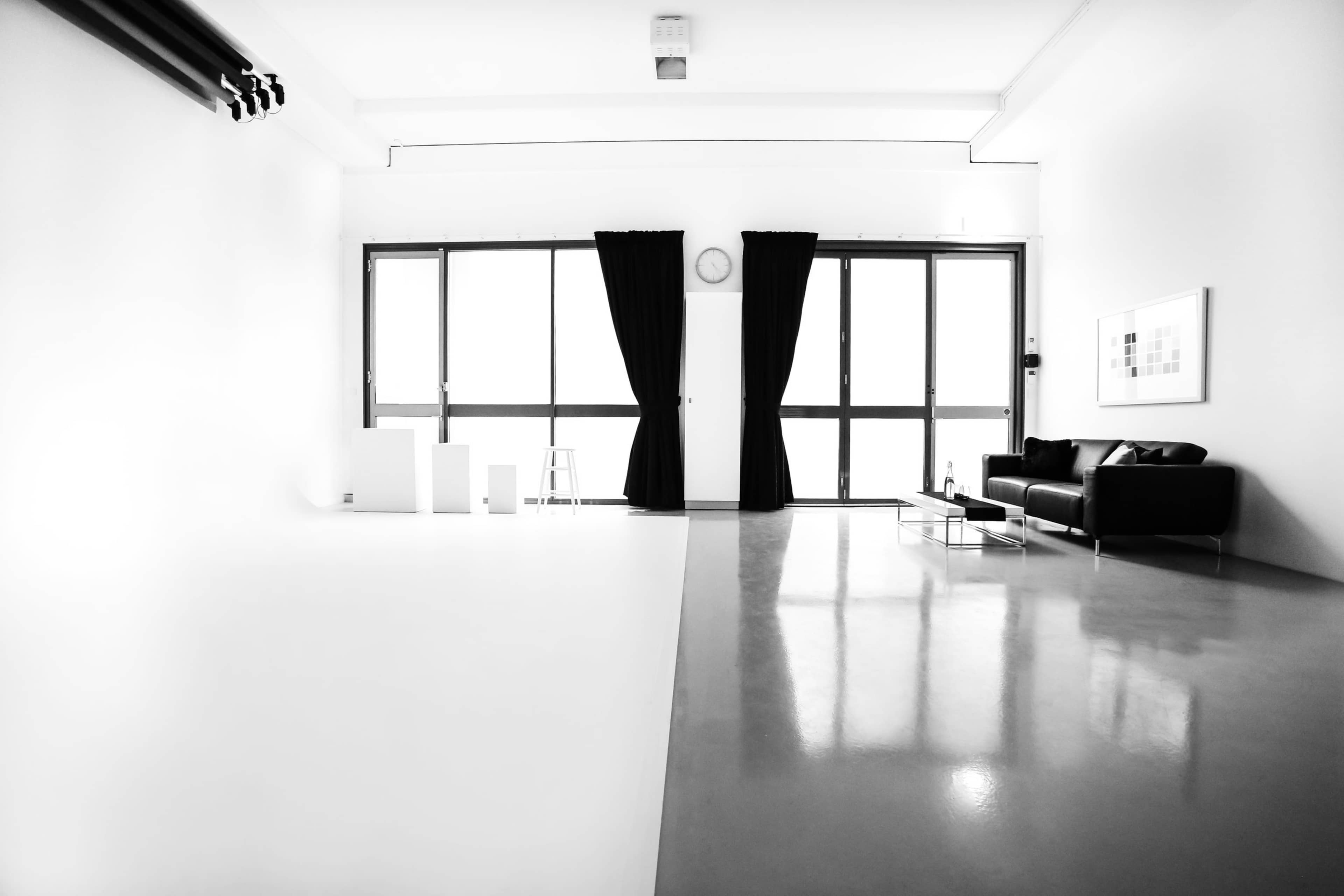 A modern room divided by a polished floor, featuring large windows with black curtains on one side and minimalistic furniture on the other.