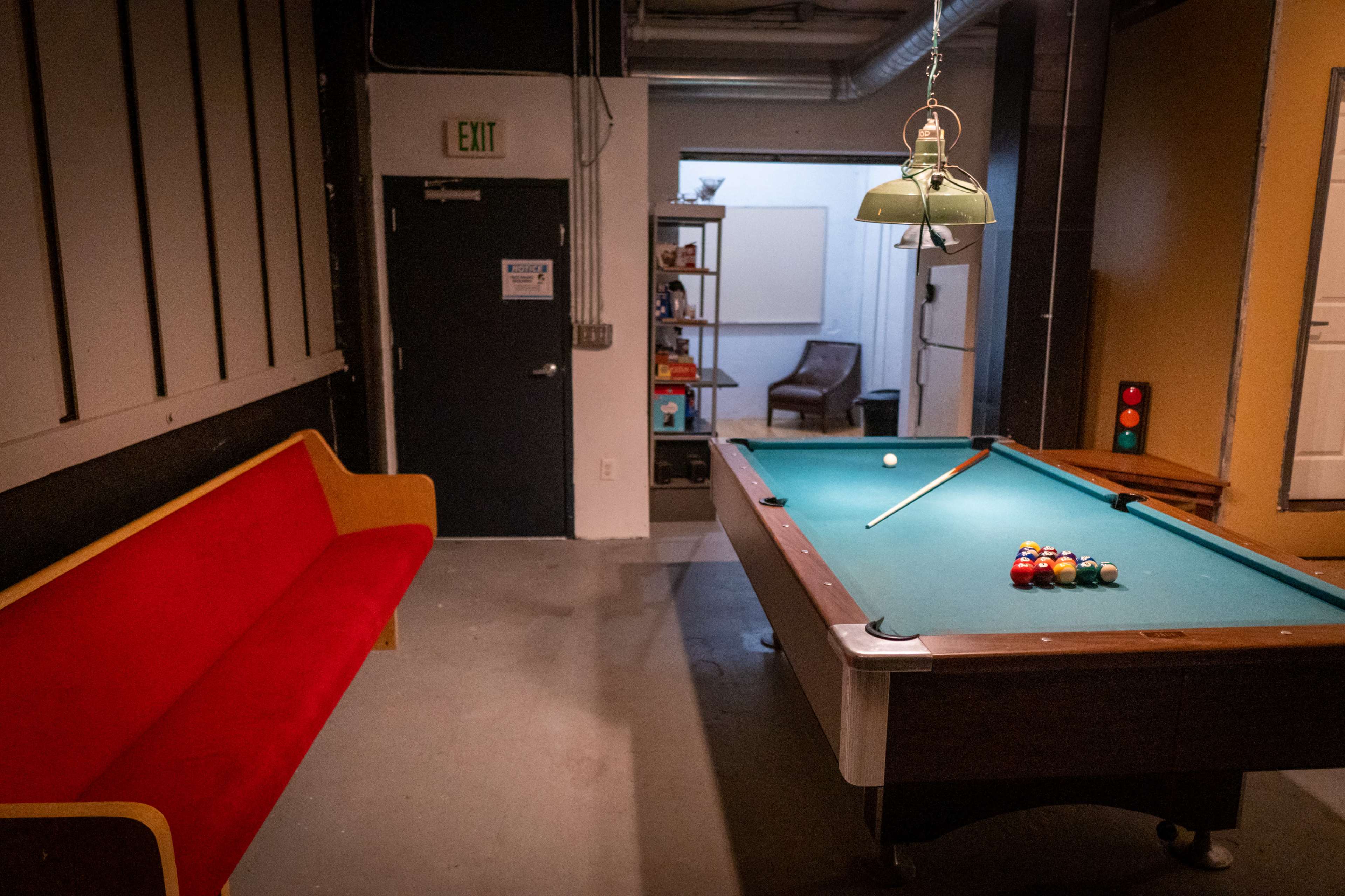 A pool table with a set of billiard balls arranged in a triangle, next to a red sofa and a door leading to another room.