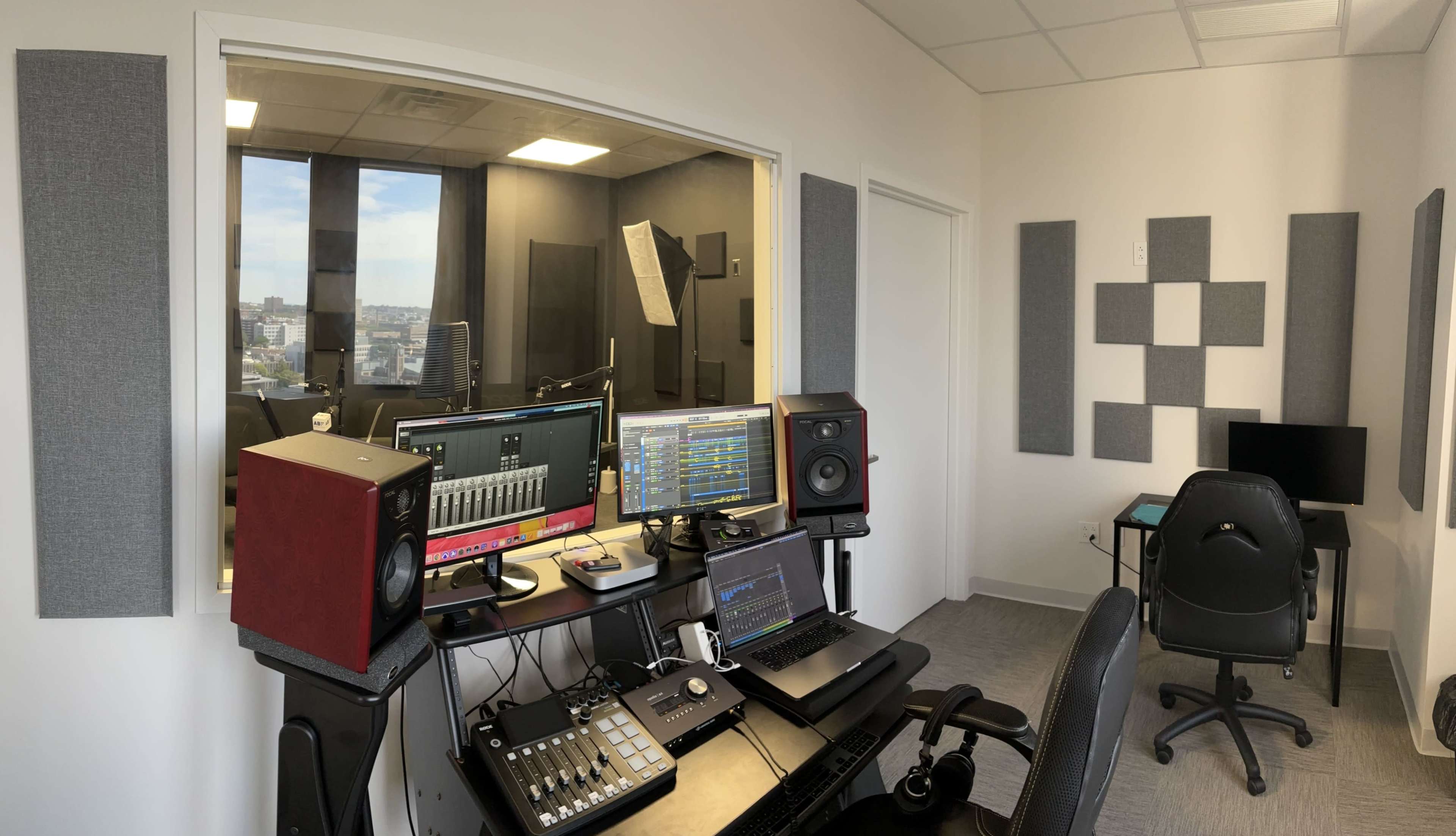 A recording studio with a desk featuring audio equipment, a laptop, and speakers, along with soundproofing panels on the walls and a window providing a view of the outside.