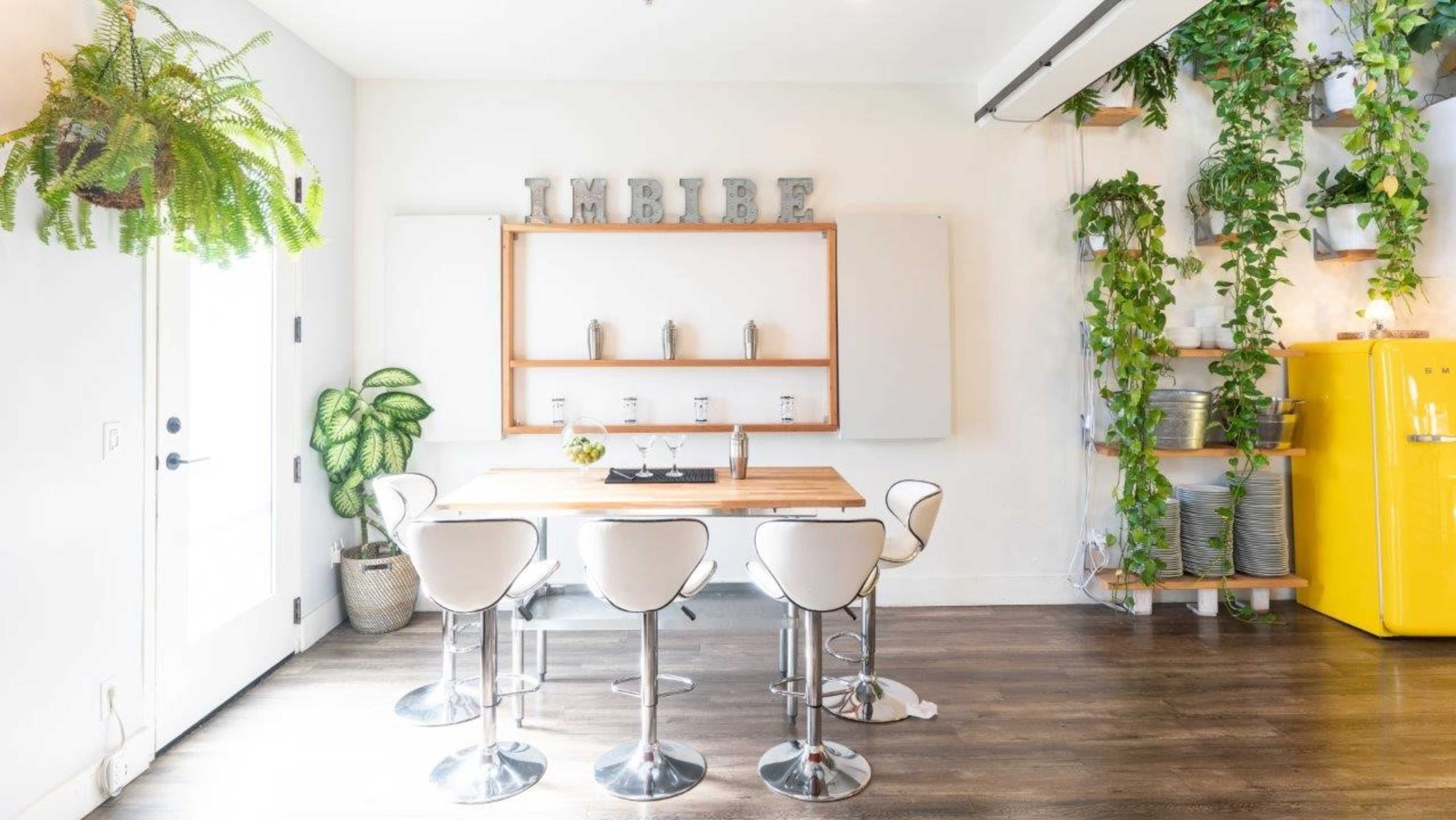 A bright kitchen features a wooden table surrounded by white bar stools, with plants and a vintage yellow refrigerator in the background.