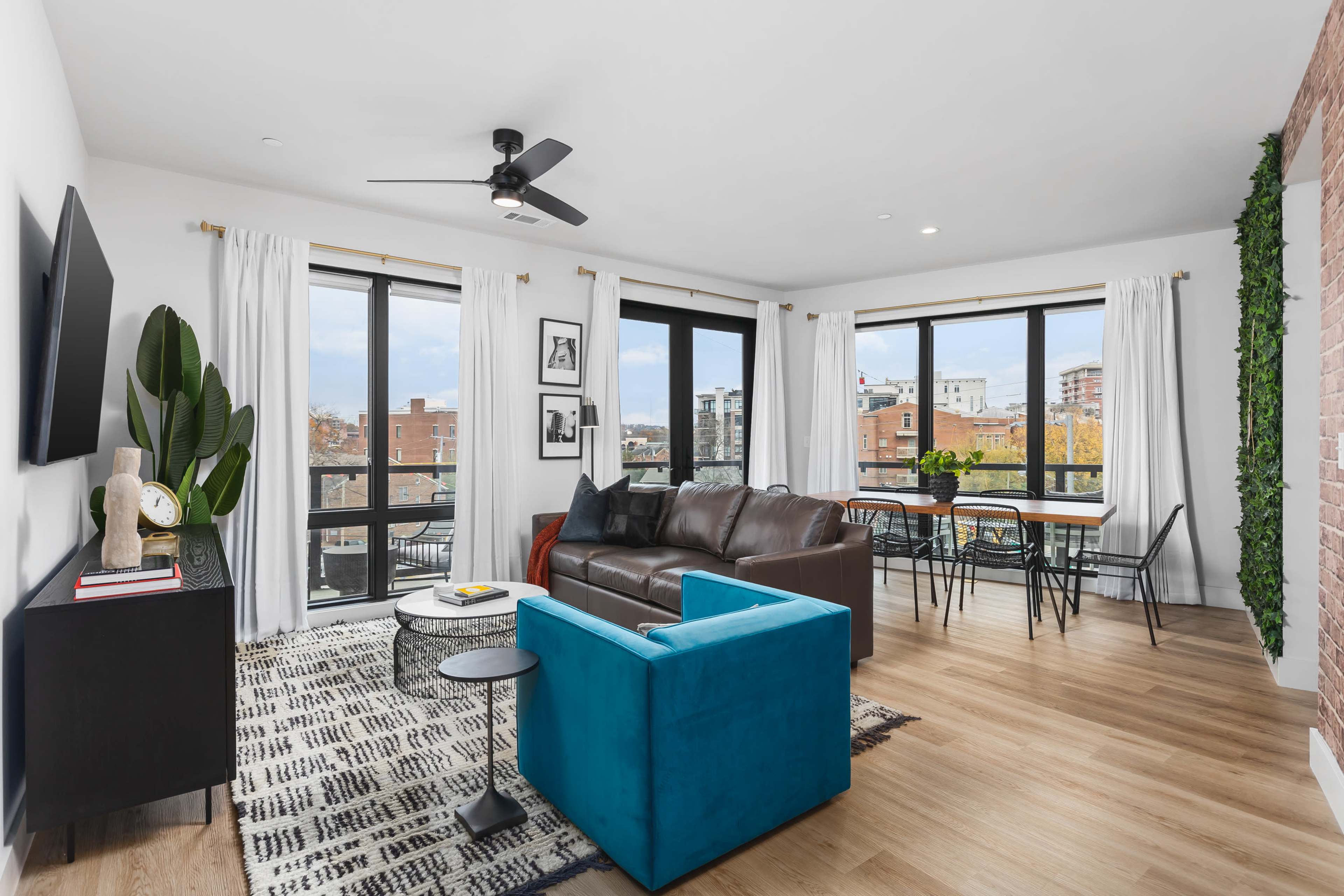 A contemporary living space features a brown leather sofa, a blue accent chair, and large windows overlooking an urban landscape.
