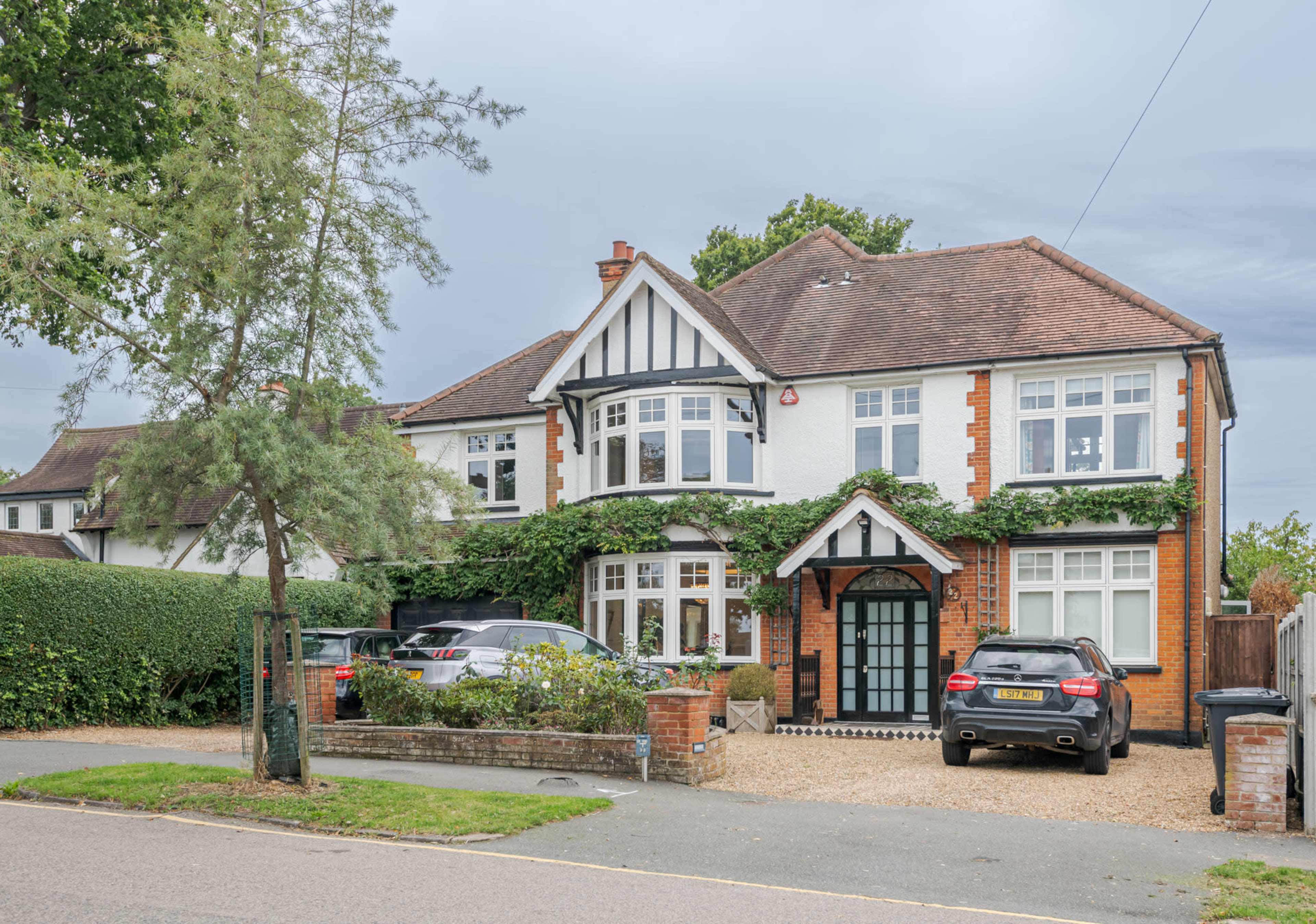 A two-story house with a gabled roof and decorative white-and-black trim is situated on a tree-lined street, surrounded by greenery and featuring a gravel driveway.