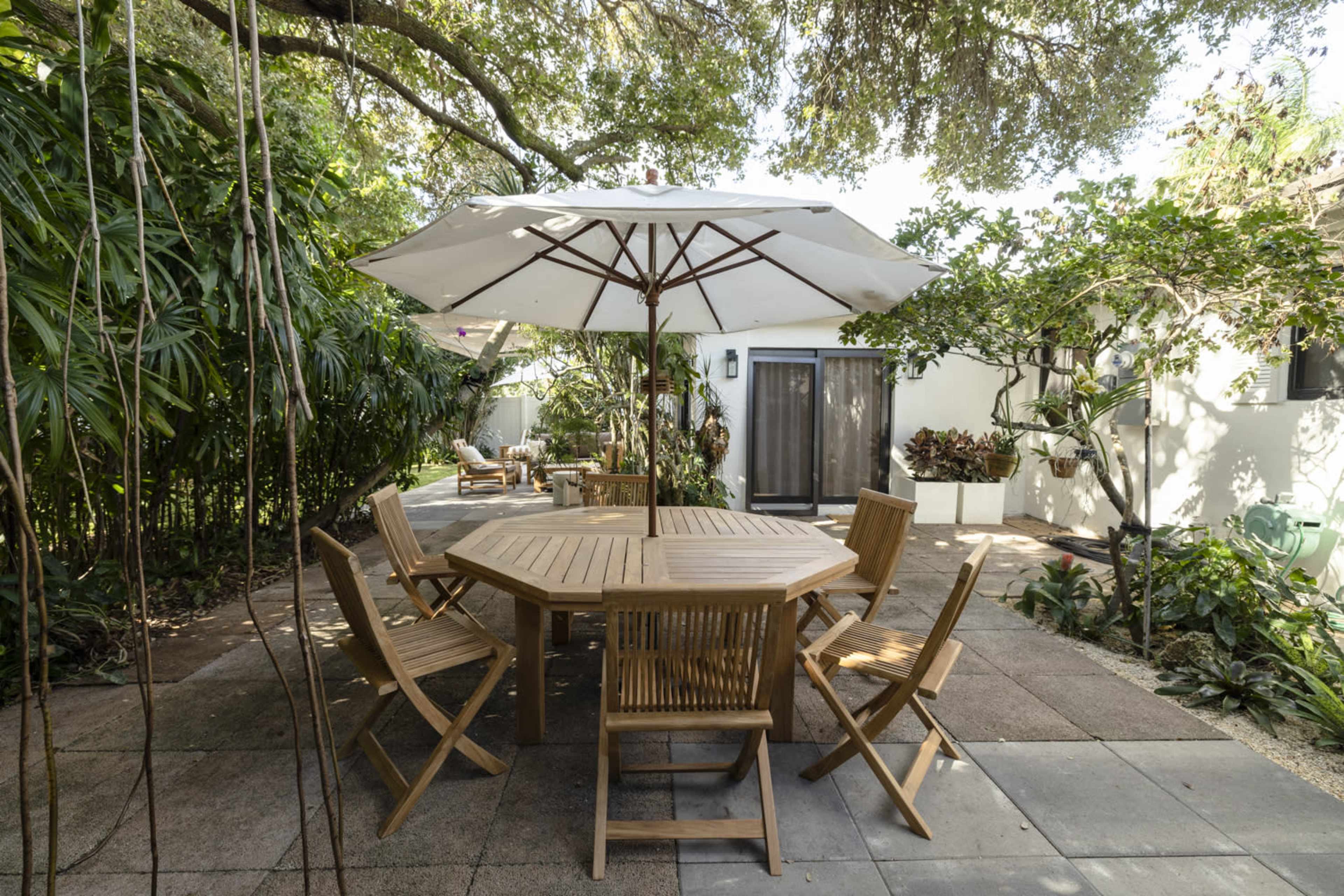 A wooden outdoor dining table is surrounded by six folding chairs under a large umbrella in a garden setting with greenery and a white building in the background.