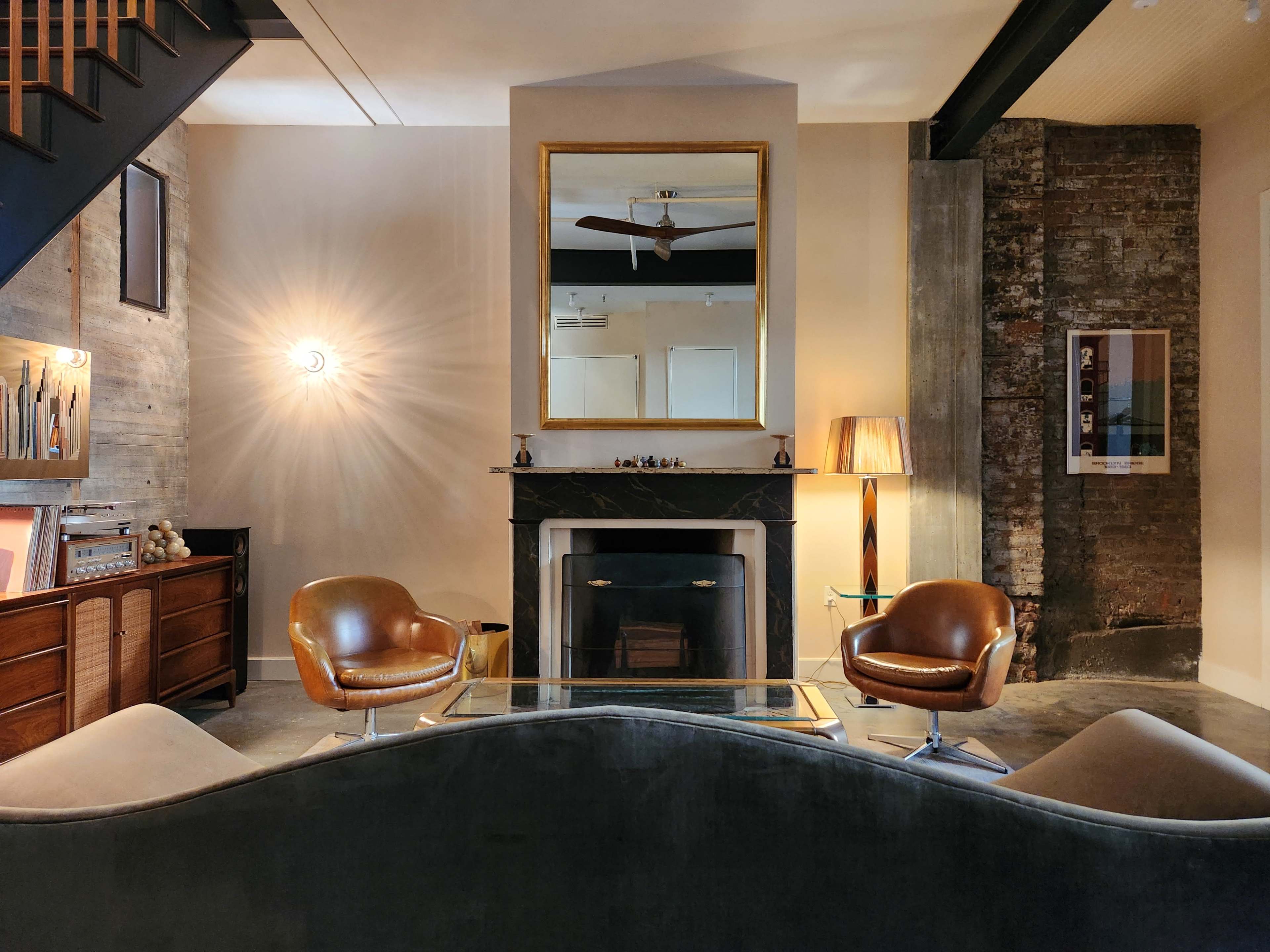 The image shows a modern living room featuring two leather chairs facing a coffee table, with a fireplace and a large mirror on the wall behind them.