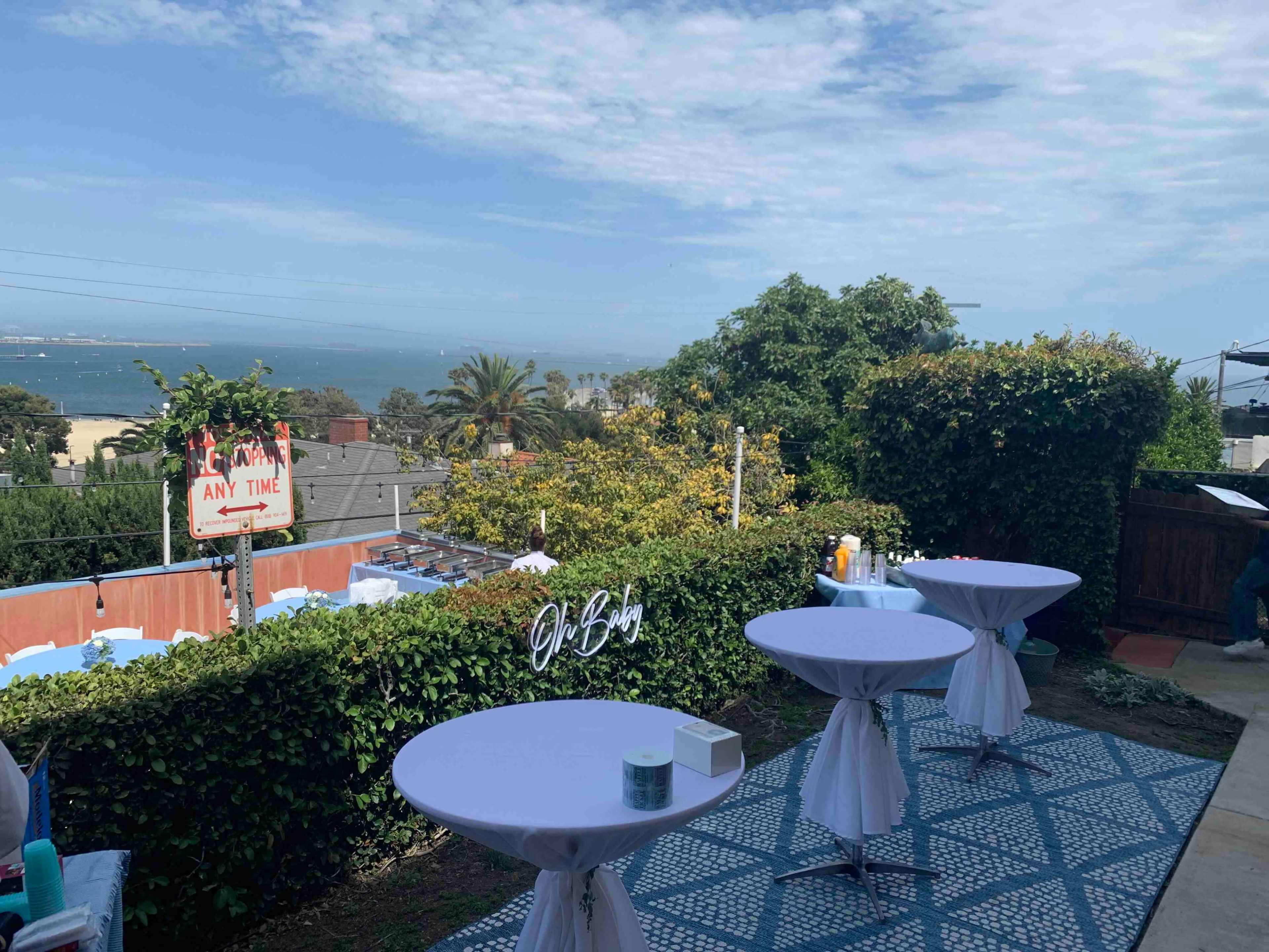 The image shows a garden setting with several round tables and a view of the ocean in the background.