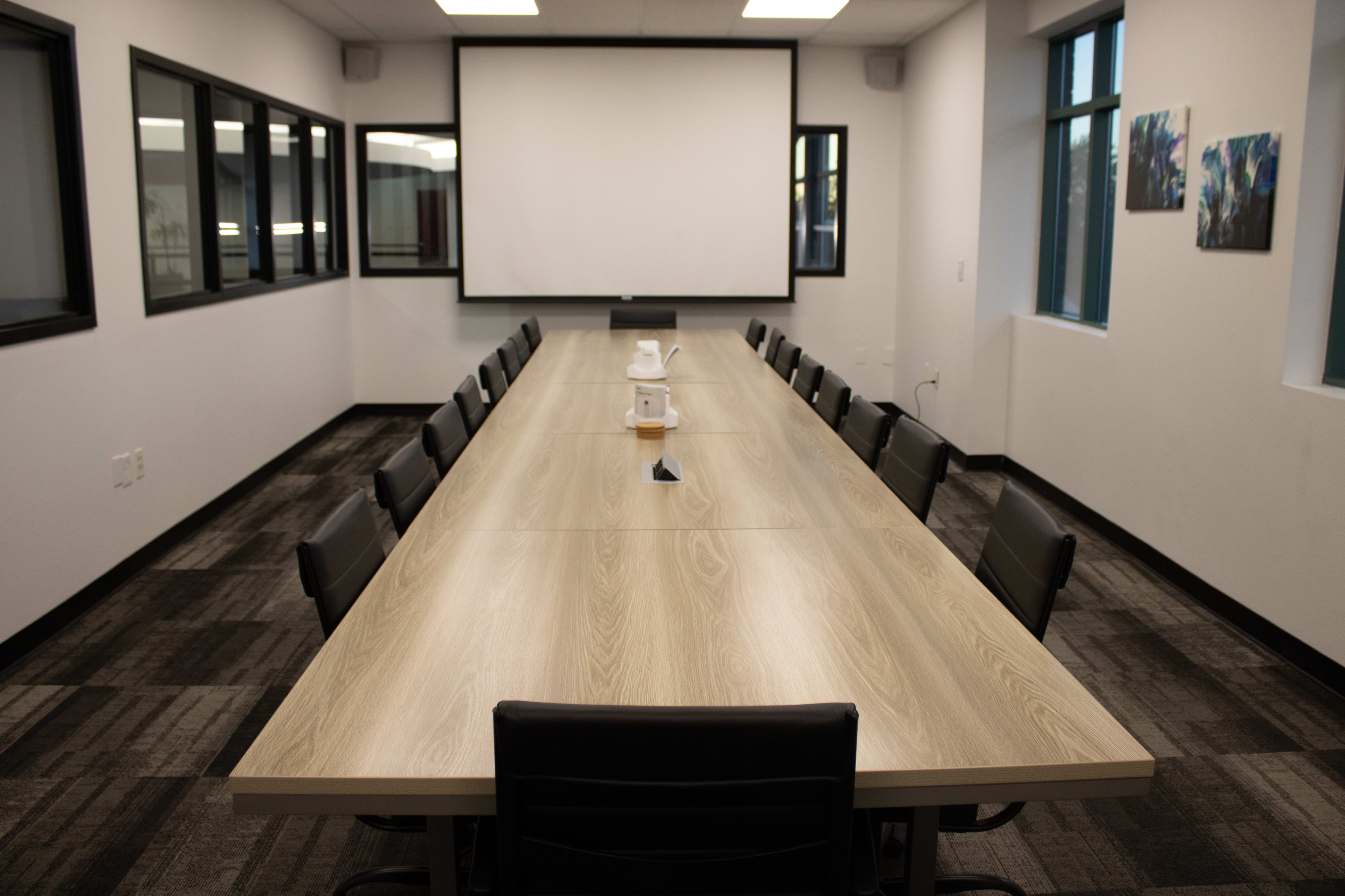 A long conference table with black chairs is set up in a brightly lit meeting room featuring a large screen at one end and windows along the walls.