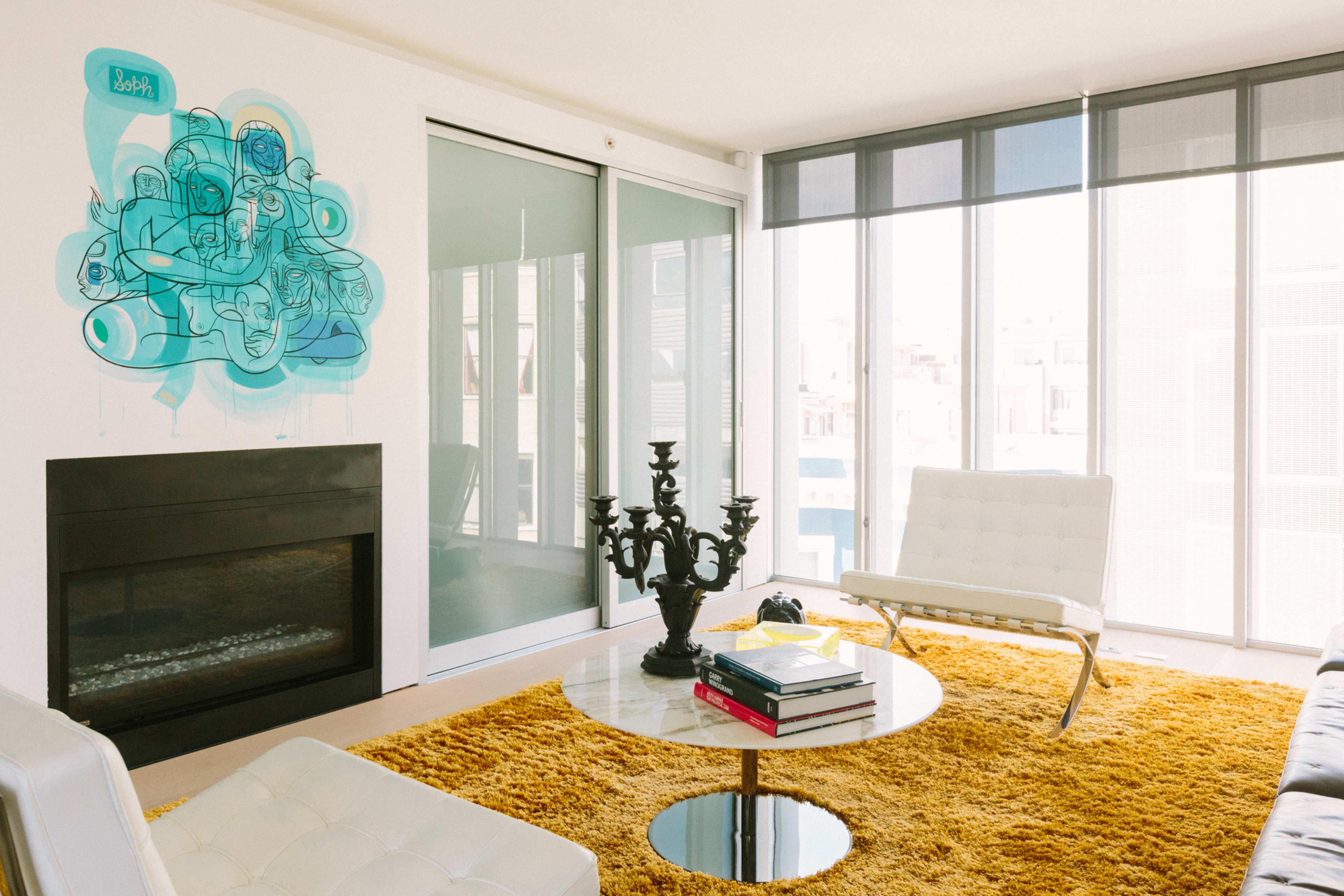 A modern living room features a white sofa, a circular glass coffee table on a yellow area rug, a black candelabra, and large windows allowing natural light to enter.