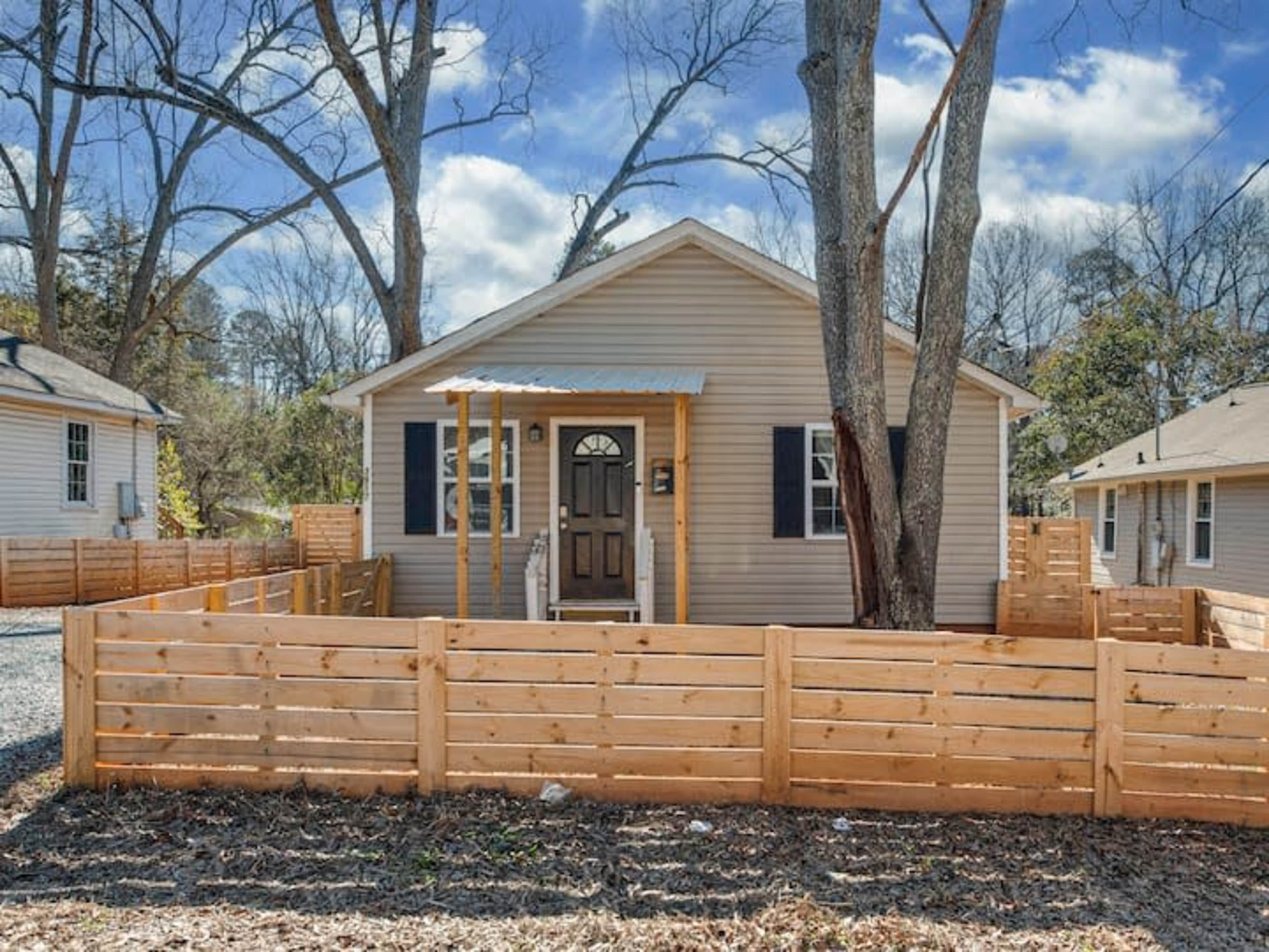 A small, single-story house with a front porch is surrounded by a wooden fence and trees.