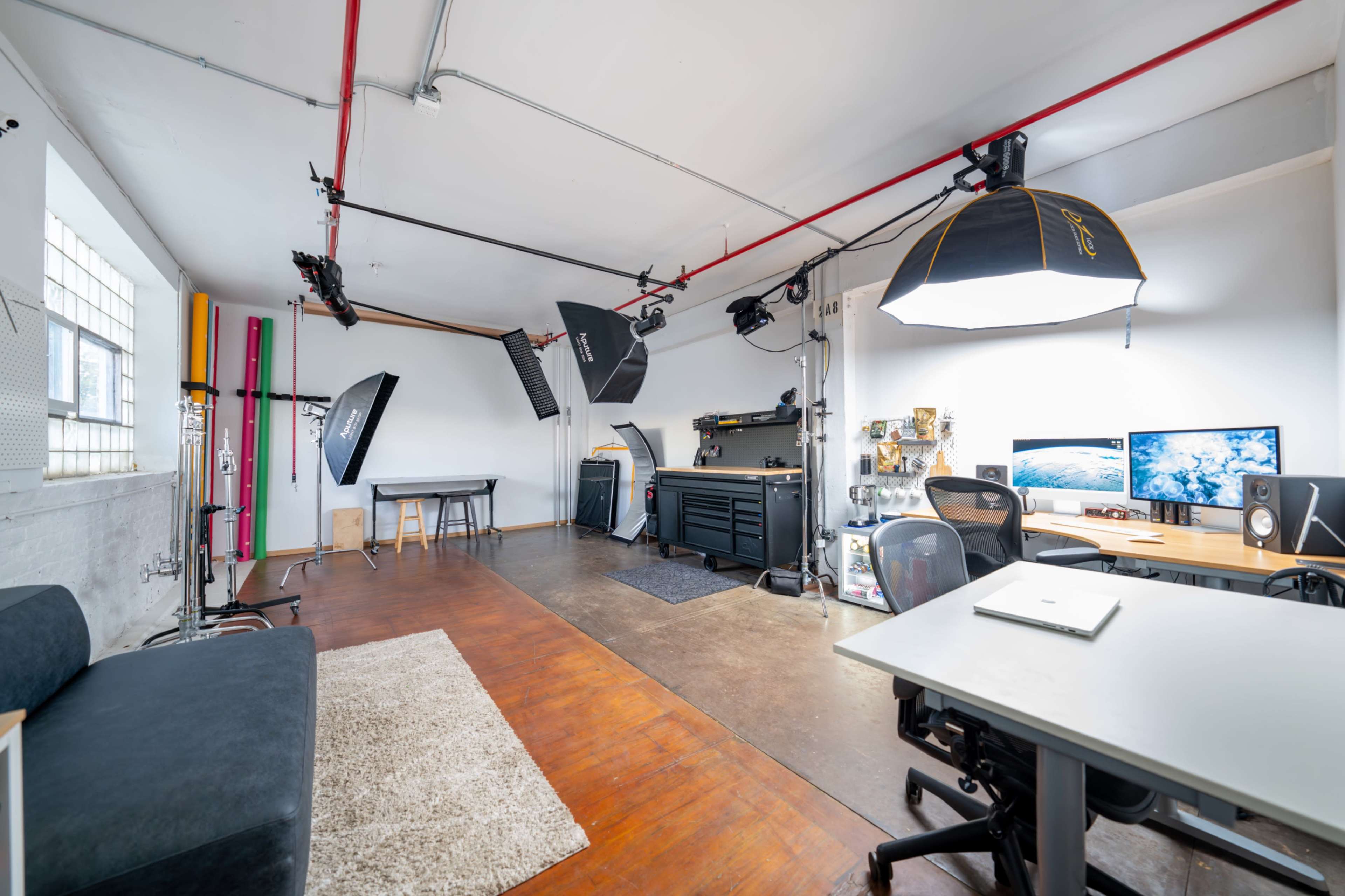 The image shows a well-equipped photography studio featuring a workspace, multiple light setups, and two computer monitors.