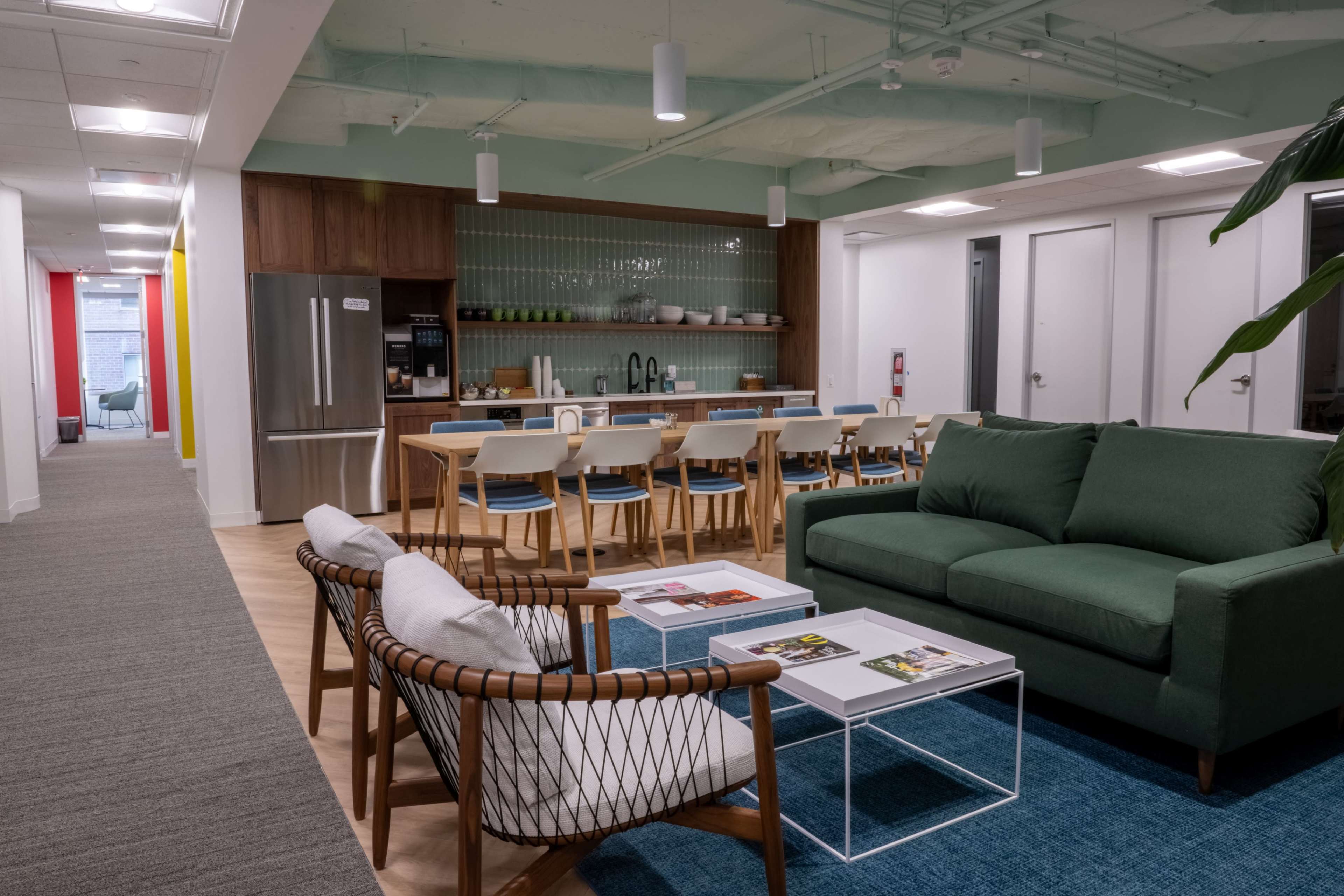 A modern office break room featuring a dining area with a long table, a kitchen with stainless steel appliances, and a lounge area with a green sofa and woven chairs.