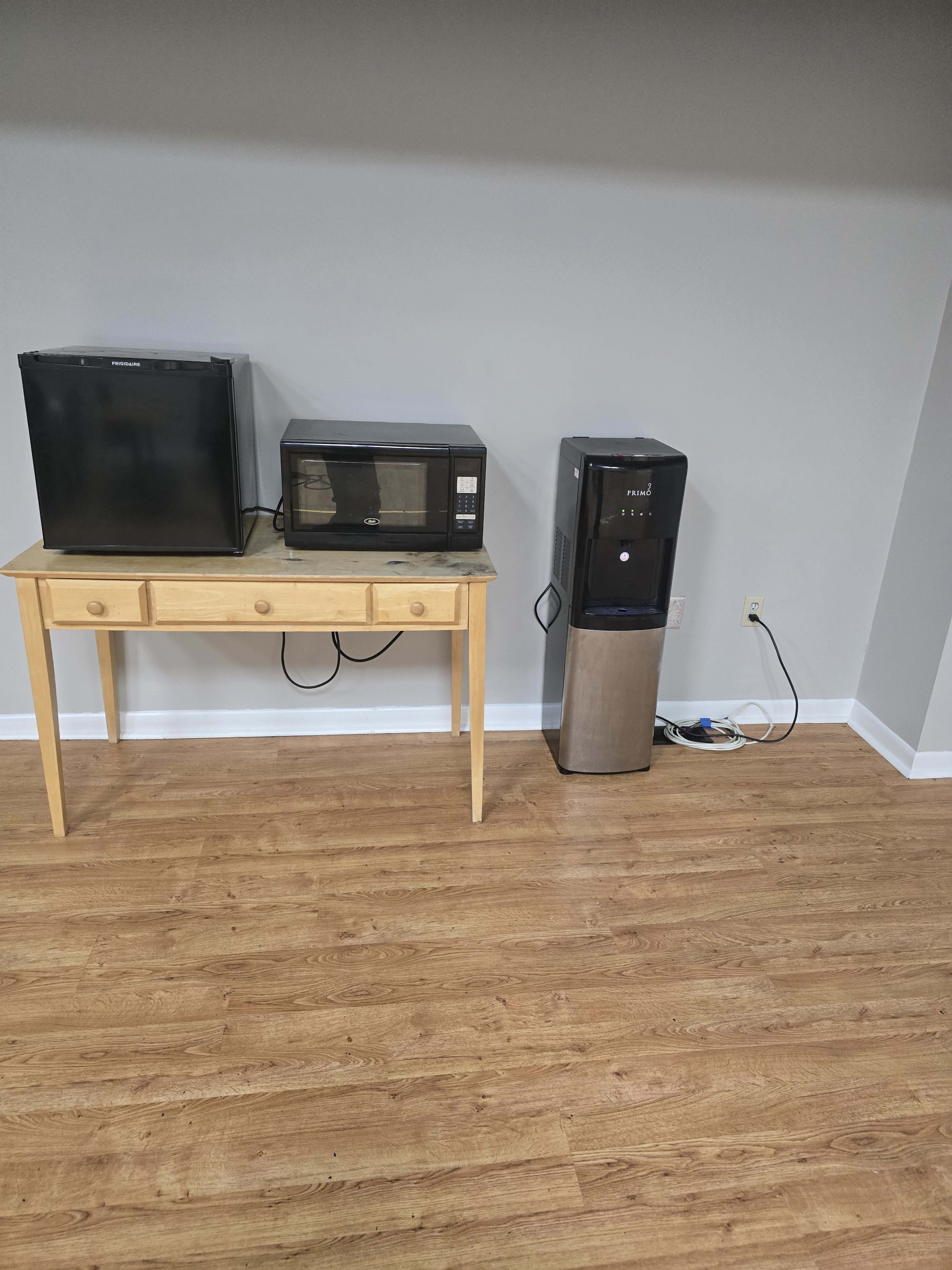 The image shows a small wooden table with a microwave and a mini fridge on top, next to a water cooler against a plain wall.