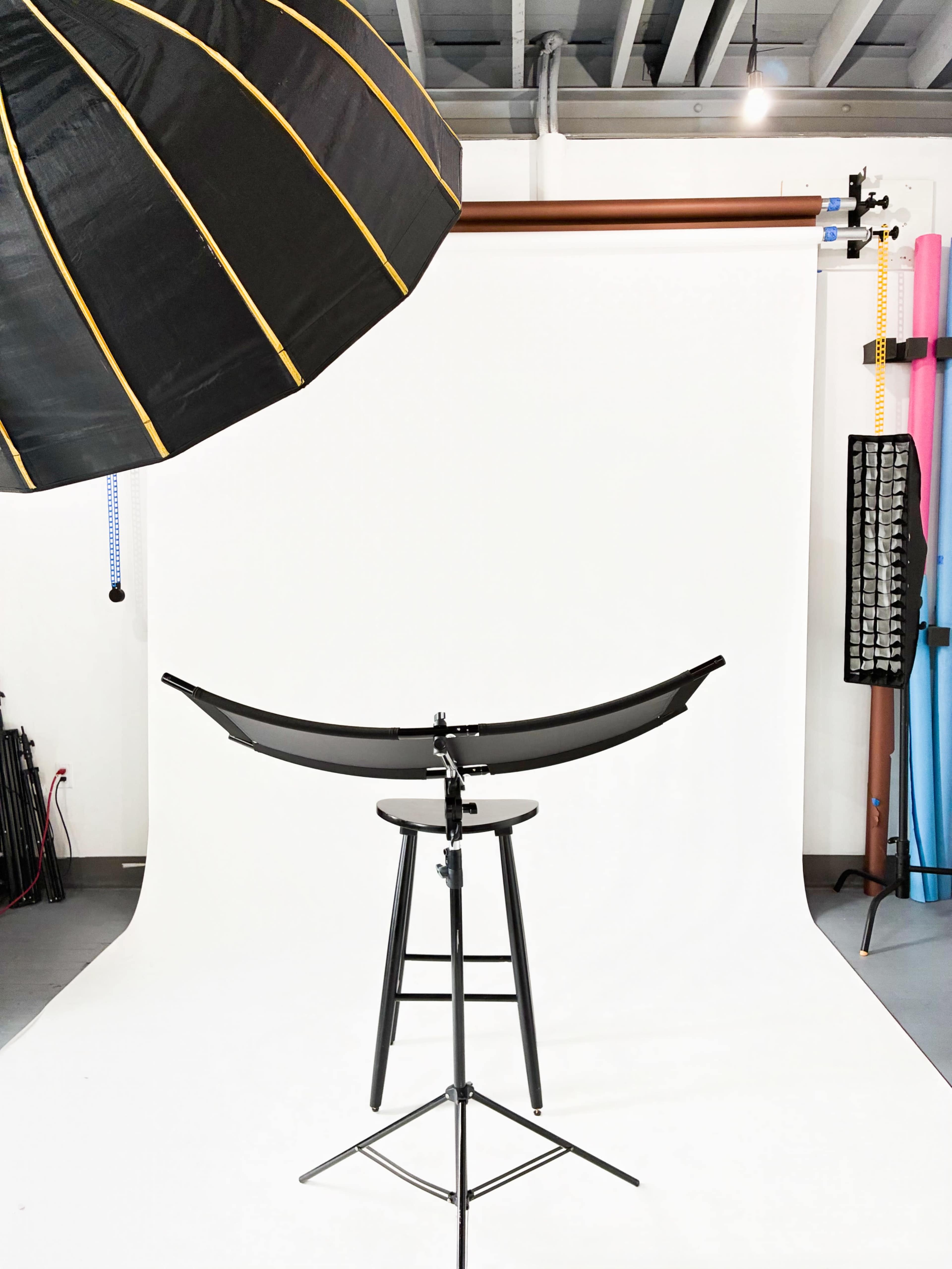 The image shows a photography setup featuring a black and gold umbrella softbox positioned above a black stool in front of a white backdrop.