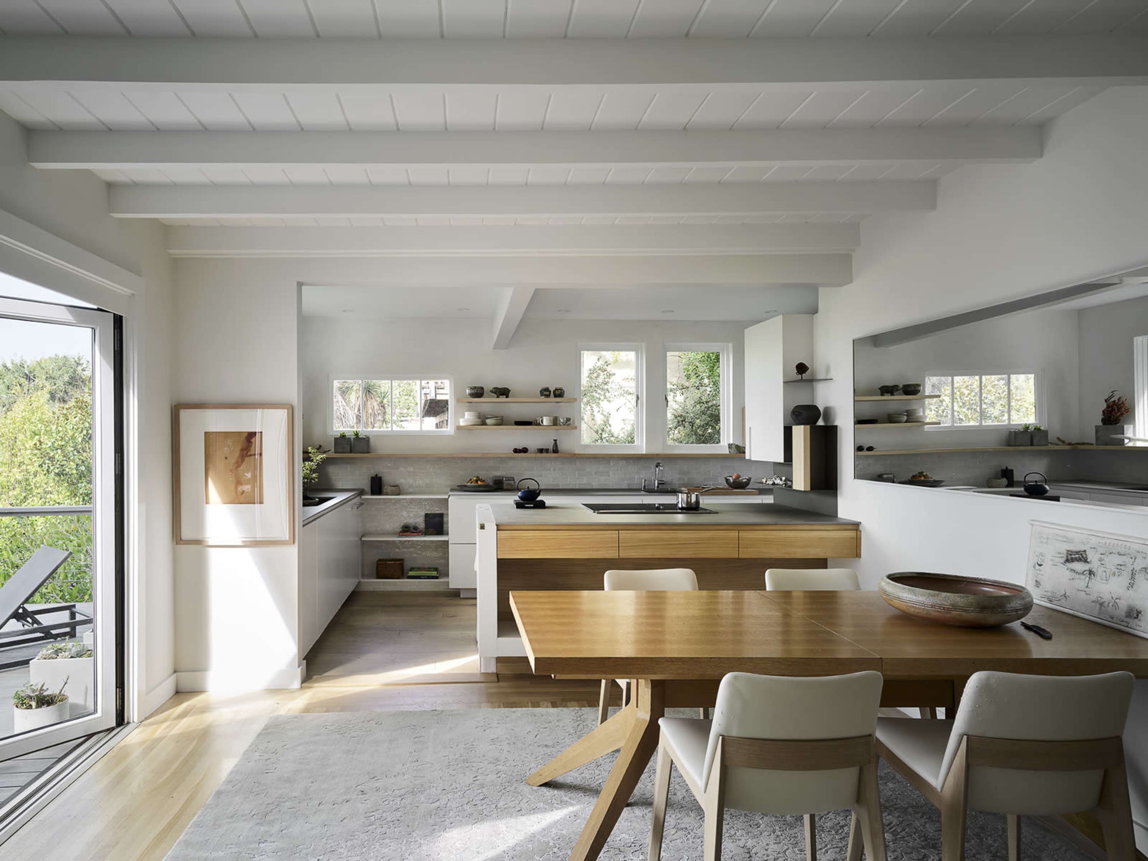 A modern kitchen features light wood beams, a central wooden dining table, and large windows that provide a view of greenery outside.