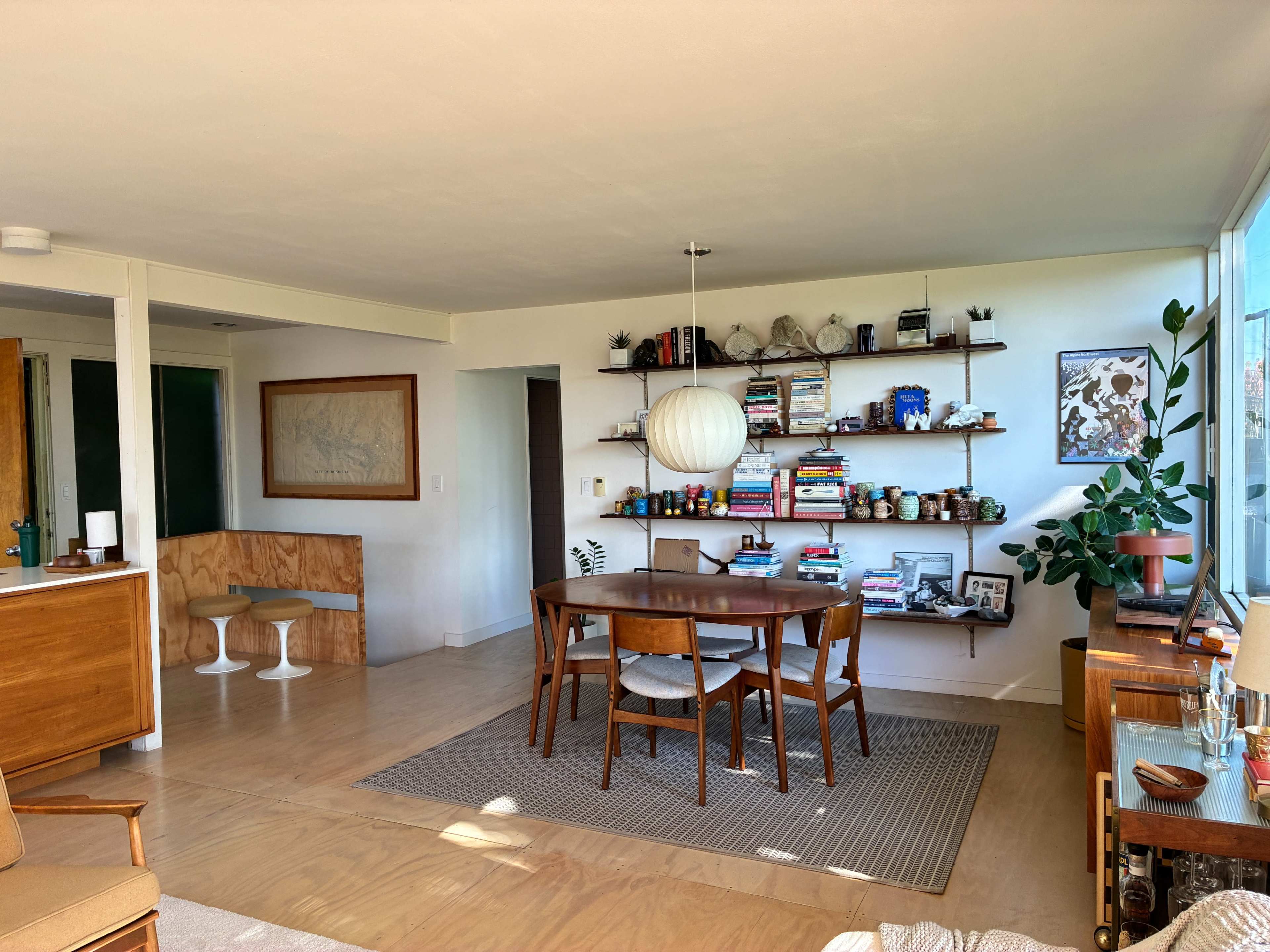 The image shows a modern living space featuring a dining area with a round wooden table, chairs, and a wall shelf filled with books and decorative items.